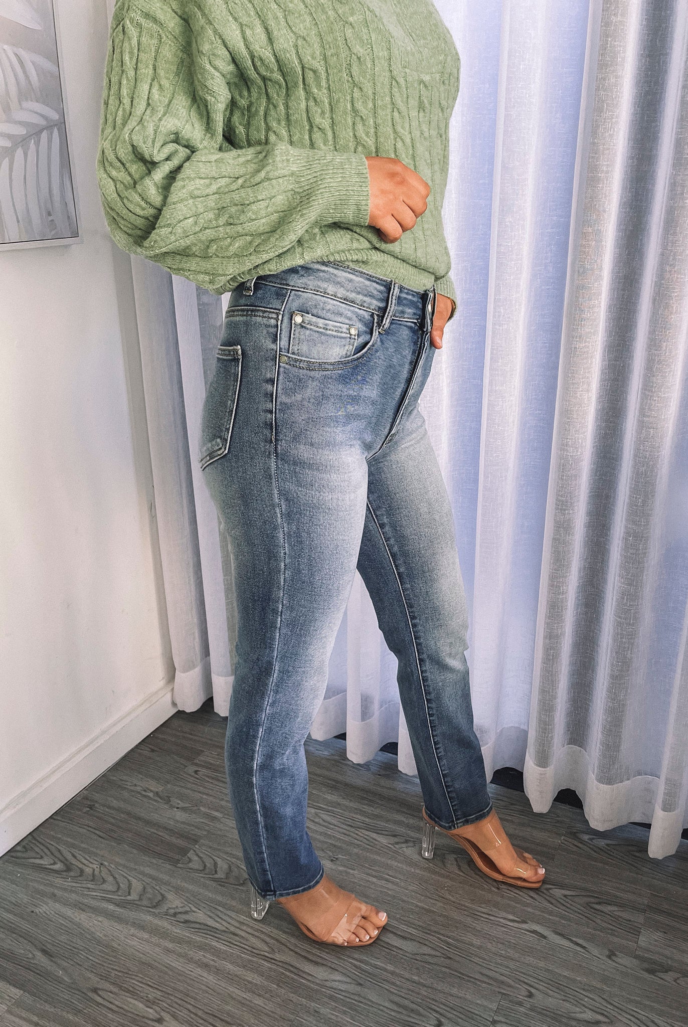 A person wearing mid-wash blue denim jeans and a green sweater, standing in a room with white curtains.