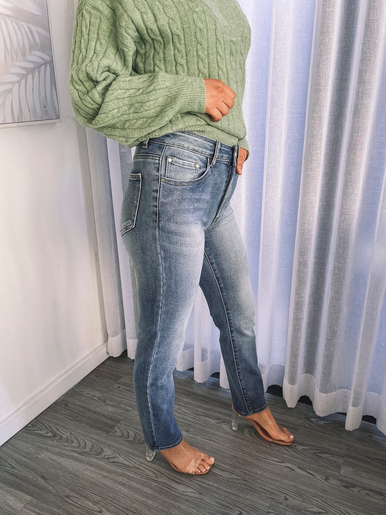 A person wearing mid-wash blue denim jeans and a green sweater, standing in a room with white curtains.