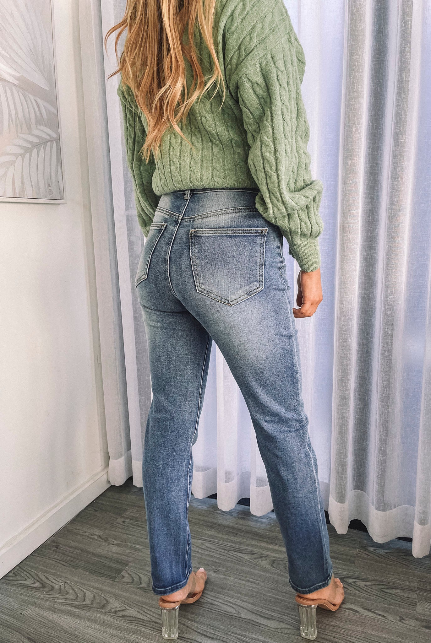 A person wearing mid-wash blue denim jeans and a green sweater, standing in a room with white curtains.