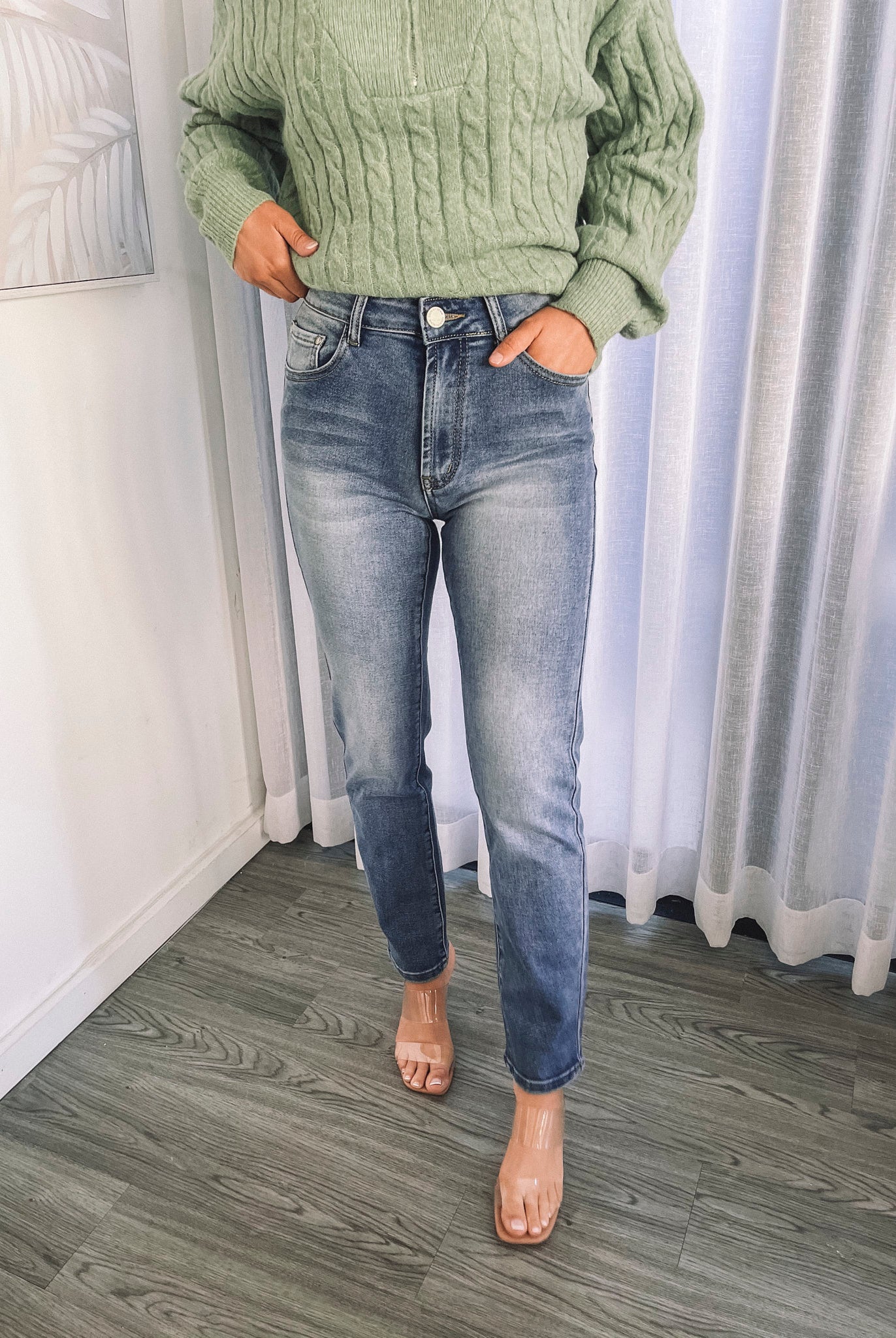A person wearing mid-wash blue denim jeans and a green sweater, standing in a room with white curtains.