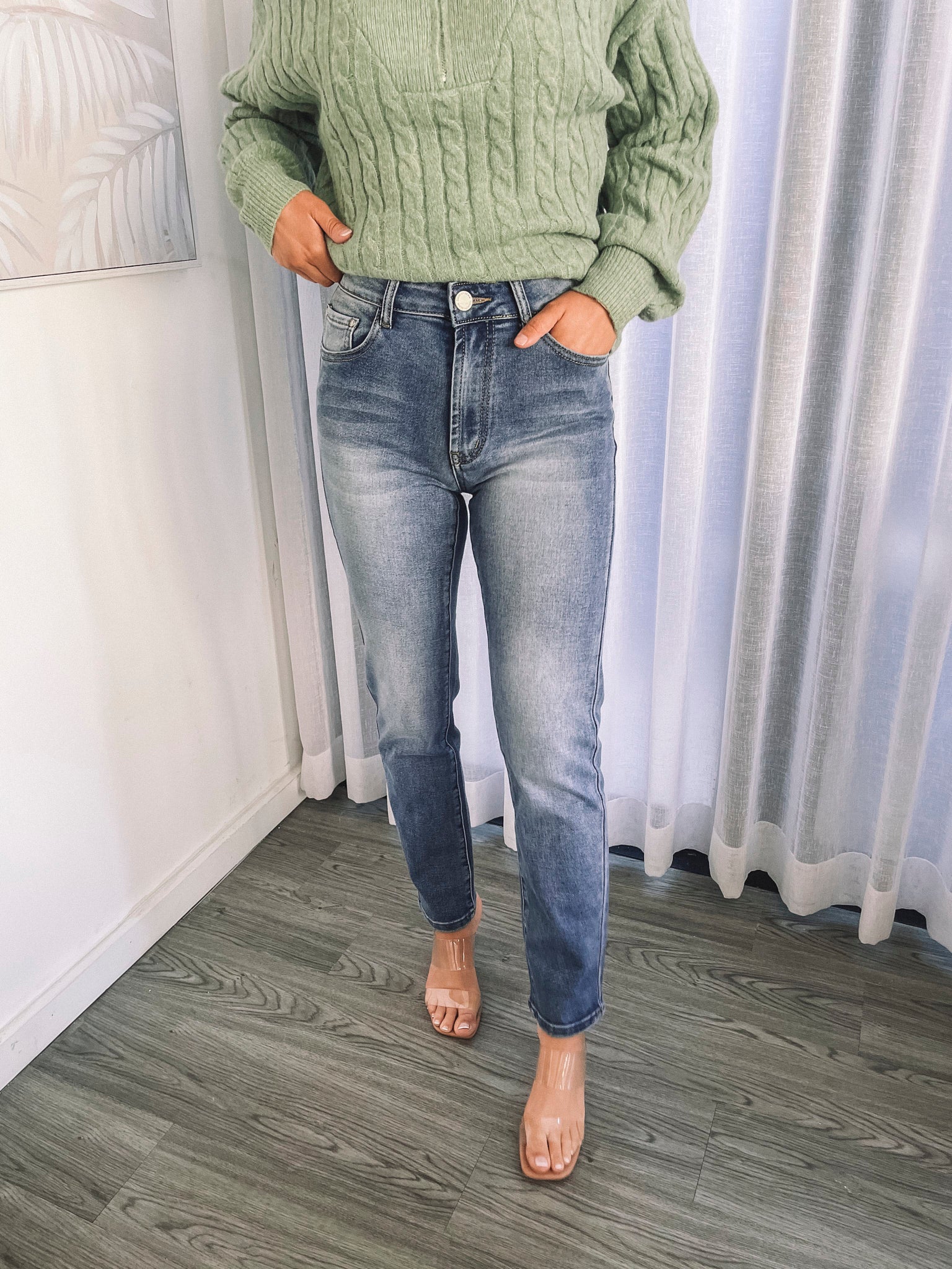 A person wearing mid-wash blue denim jeans and a green sweater, standing in a room with white curtains.