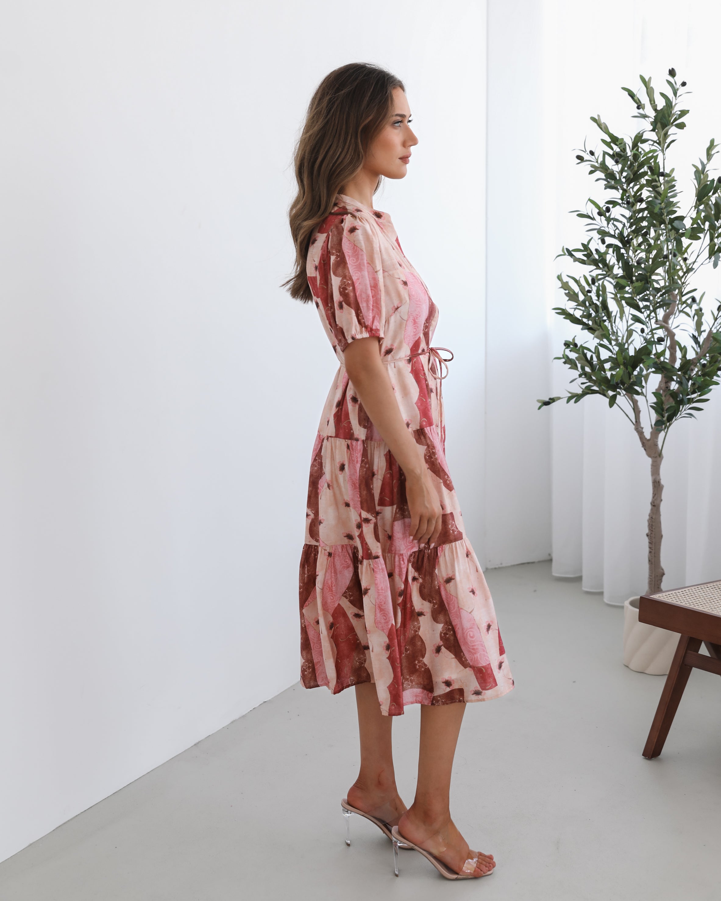 A woman standing in a room with a plant in the background, wearing a multi-toned print dress with short puff sleeves, a collar with a v-neck, center-front buttons, and a tiered skirt.