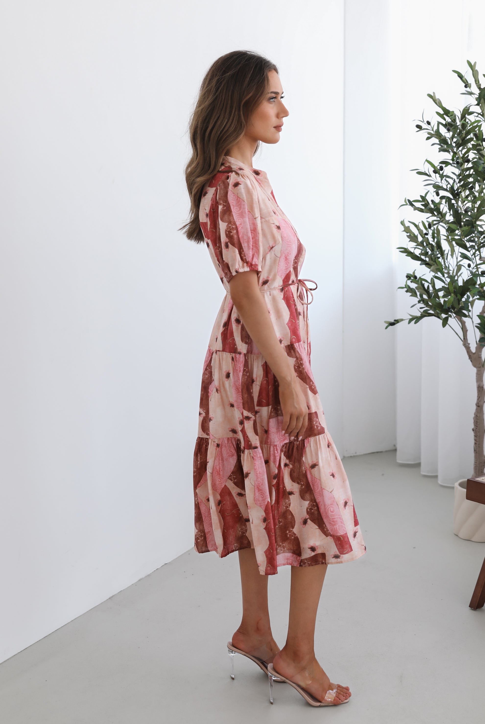 A woman standing in a room with a plant in the background, wearing a multi-toned print dress with short puff sleeves, a collar with a v-neck, center-front buttons, and a tiered skirt.