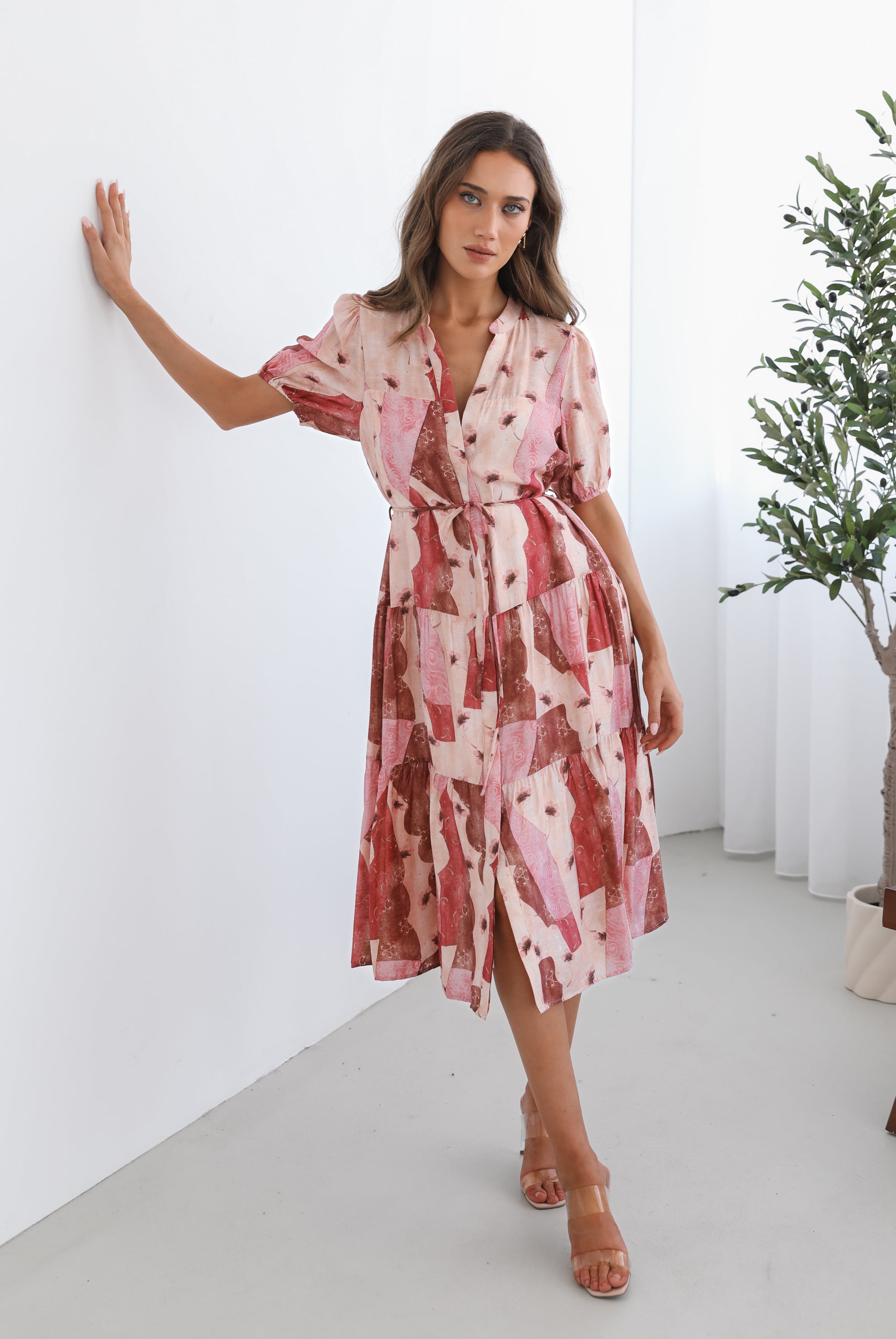A woman standing in a room with a plant in the background, wearing a multi-toned print dress with short puff sleeves, a collar with a v-neck, center-front buttons, and a tiered skirt.