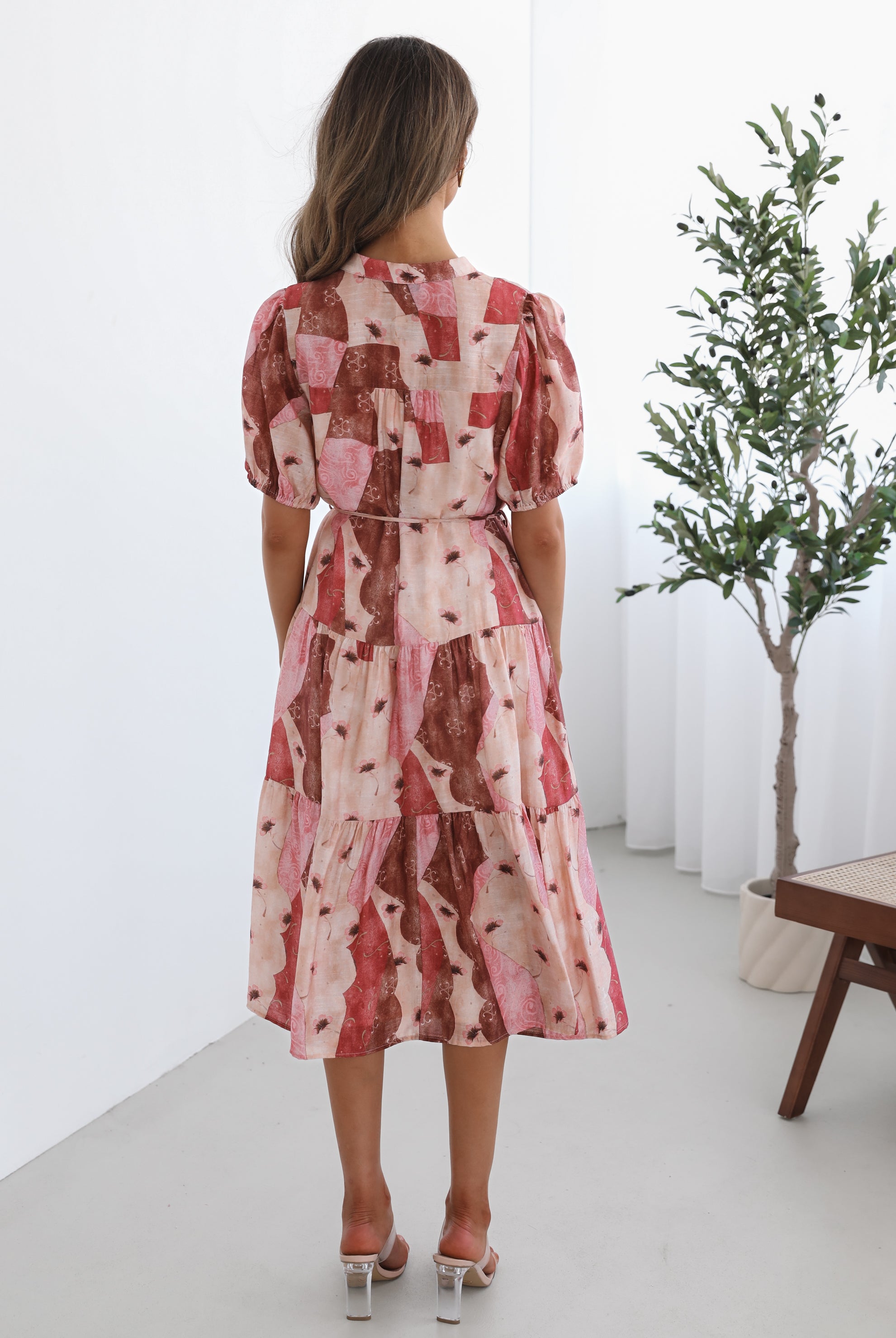A woman standing in a room with a plant in the background, wearing a multi-toned print dress with short puff sleeves, a collar with a v-neck, center-front buttons, and a tiered skirt.
