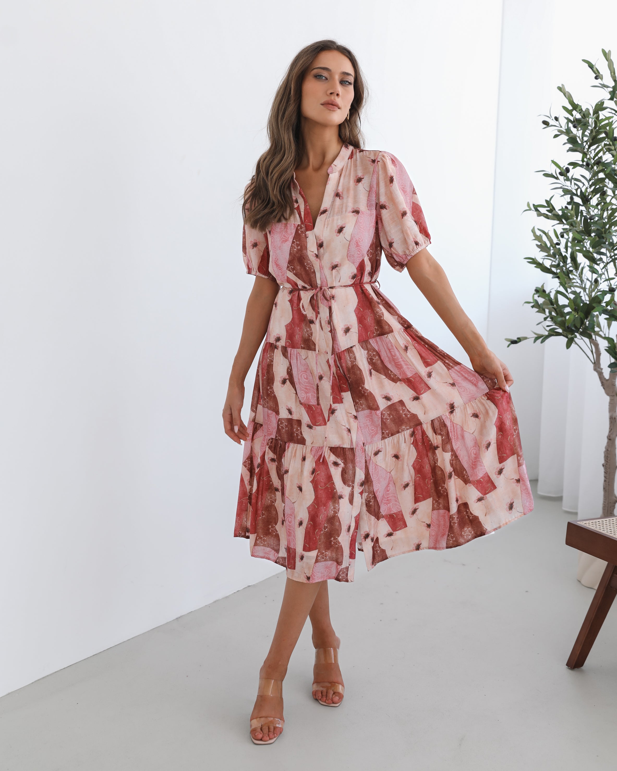 A woman standing in a room with a plant in the background, wearing a multi-toned print dress with short puff sleeves, a collar with a v-neck, center-front buttons, and a tiered skirt.