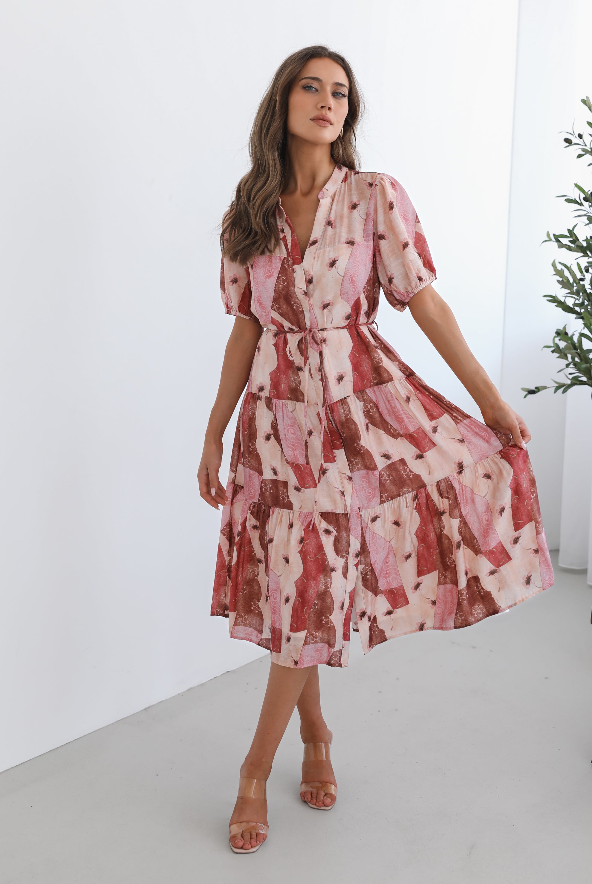 A woman standing in a room with a plant in the background, wearing a multi-toned print dress with short puff sleeves, a collar with a v-neck, center-front buttons, and a tiered skirt.