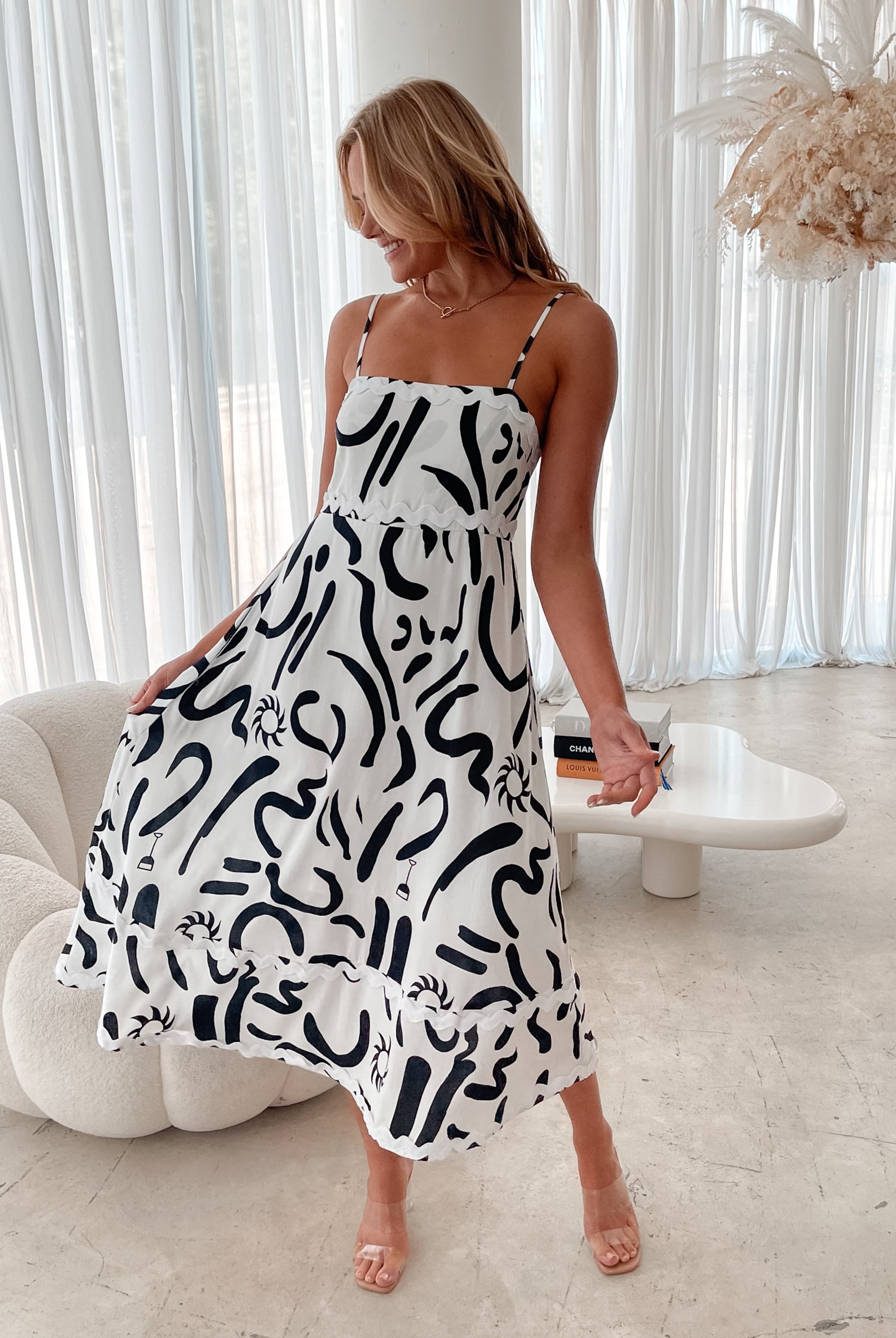 A woman standing in a room with a white dress with a black abstract print, featuring a square neckline and spaghetti straps.