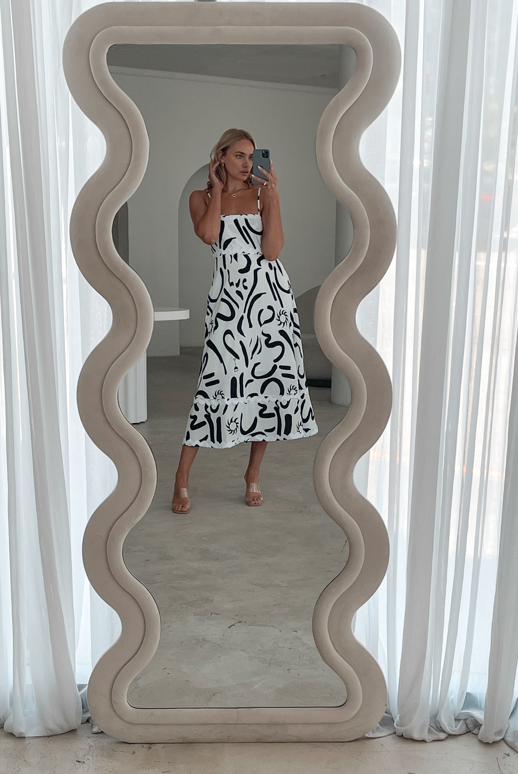A woman standing in a room with a white dress with a black abstract print, featuring a square neckline and spaghetti straps.