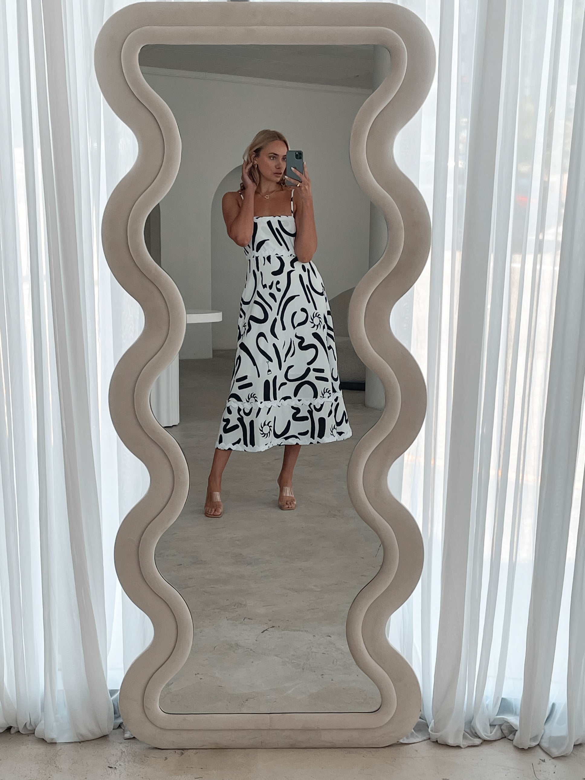 A woman standing in a room with a white dress with a black abstract print, featuring a square neckline and spaghetti straps.