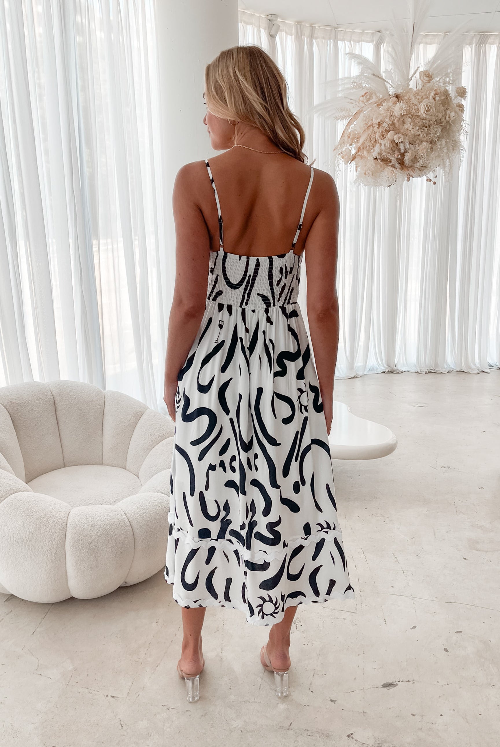 A woman standing in a room with a white dress with a black abstract print, featuring a square neckline and spaghetti straps.