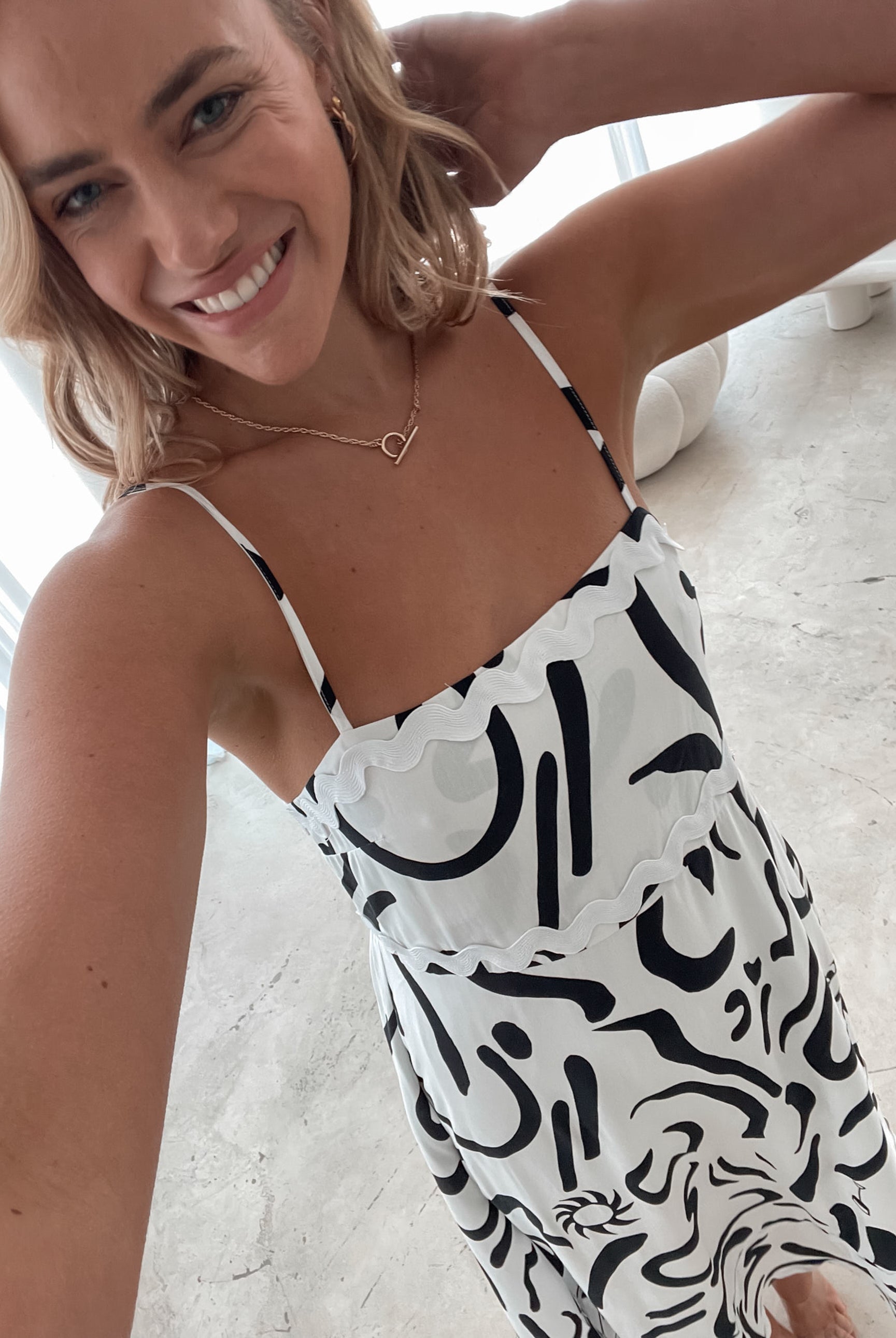 A woman standing in a room with a white dress with a black abstract print, featuring a square neckline and spaghetti straps.