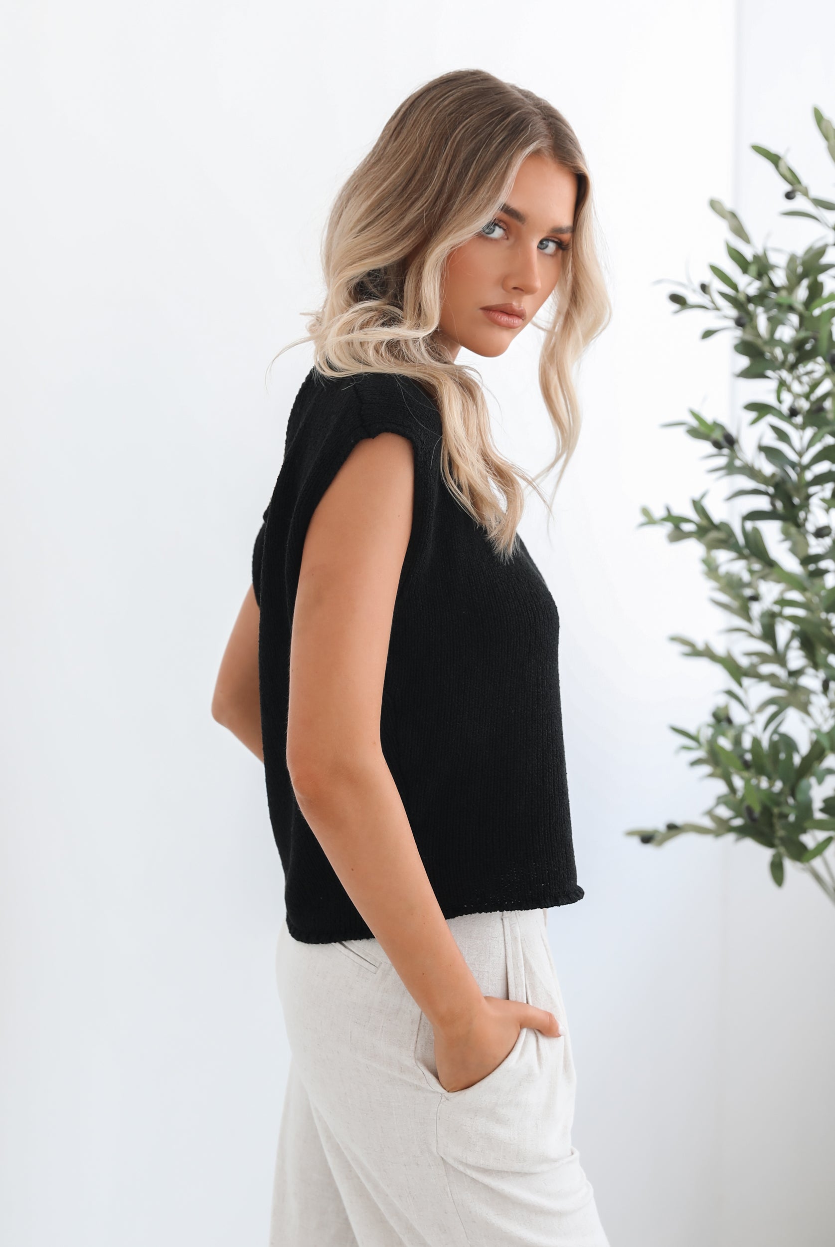 A female model wearing a black knit top with a one side off shoulder design, paired with light-colored trousers.