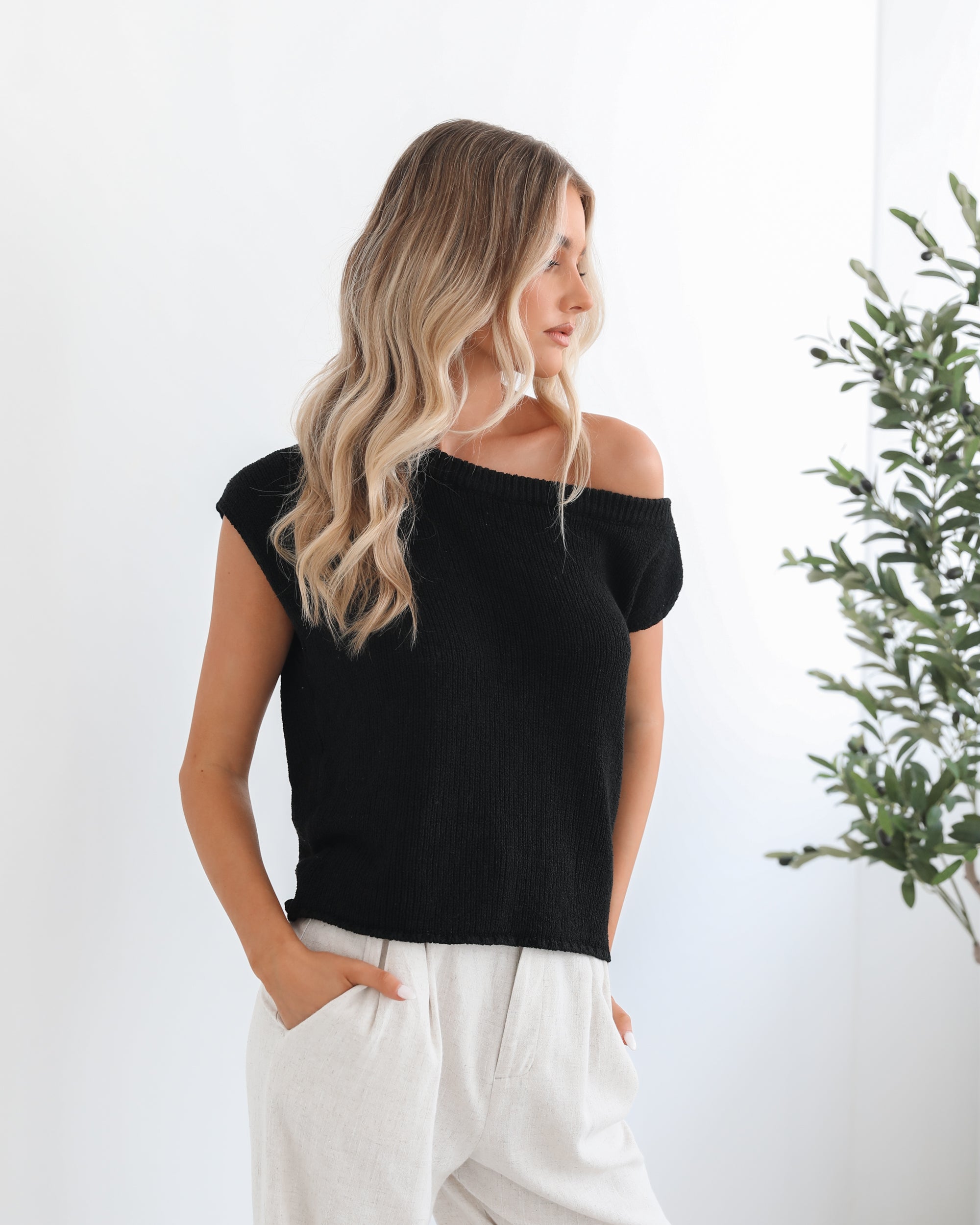 A female model wearing a black knit top with a one side off shoulder design, paired with light-colored trousers.