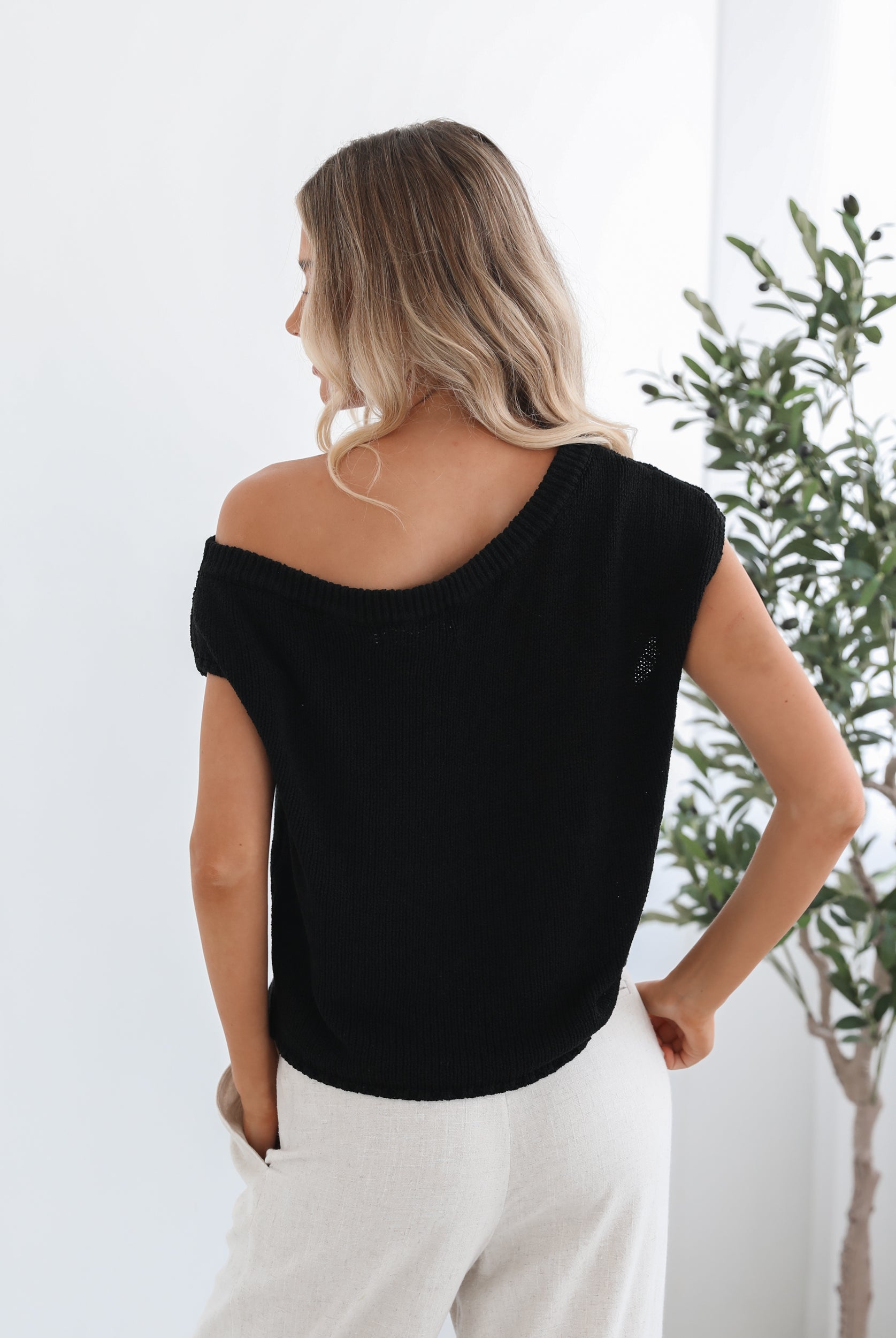 A female model wearing a black knit top with a one side off shoulder design, paired with light-colored trousers.