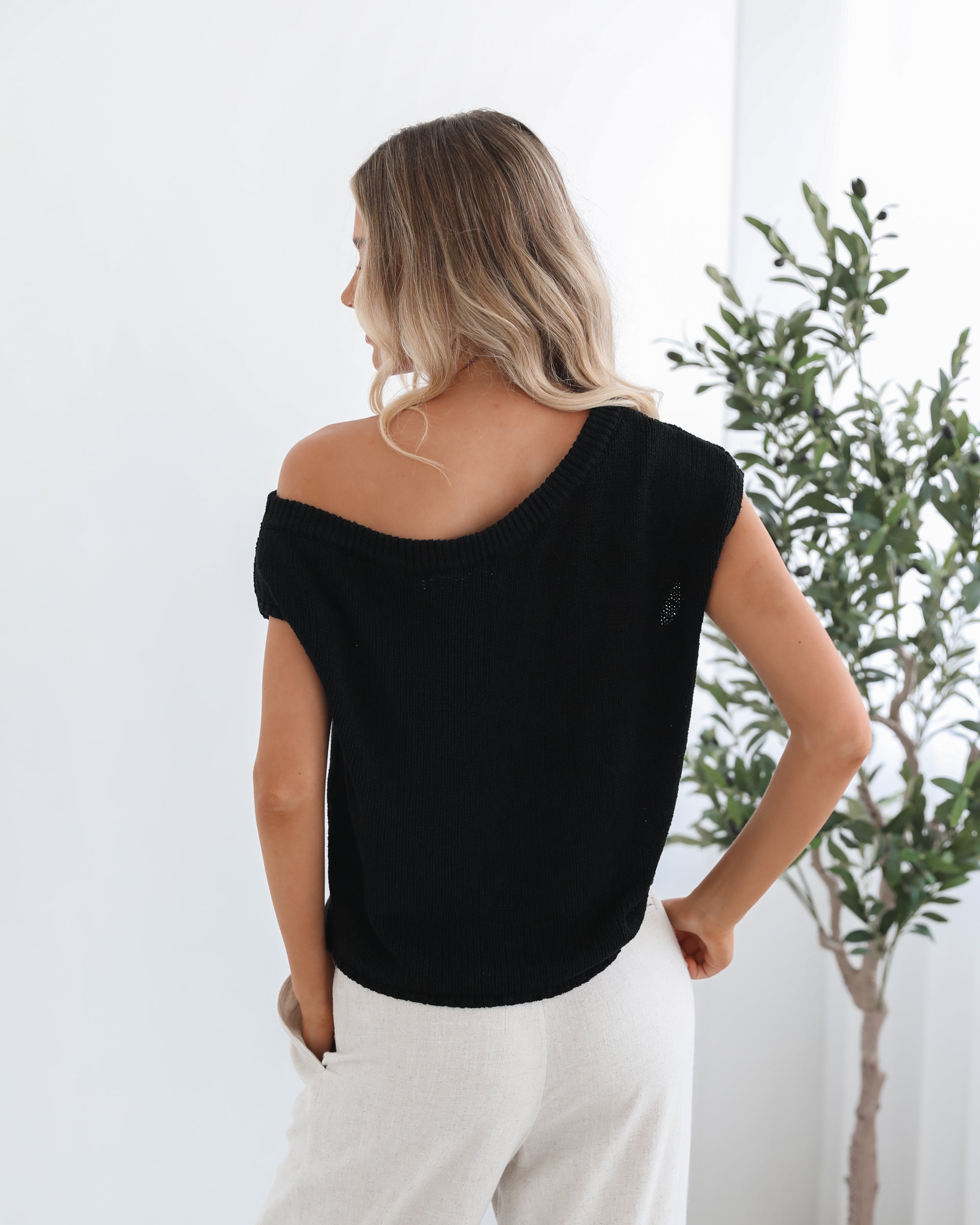 A female model wearing a black knit top with a one side off shoulder design, paired with light-colored trousers.