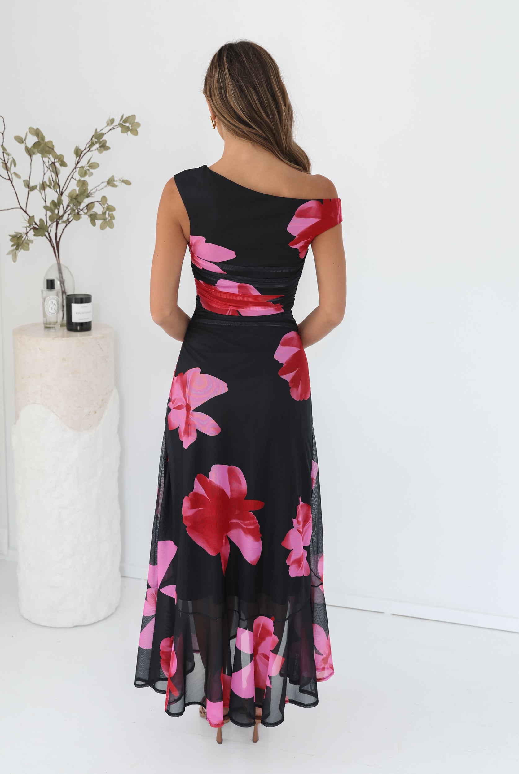 A woman standing in a white room wearing a black floral maxi dress with asymmetric neckline and ruffled skirt.