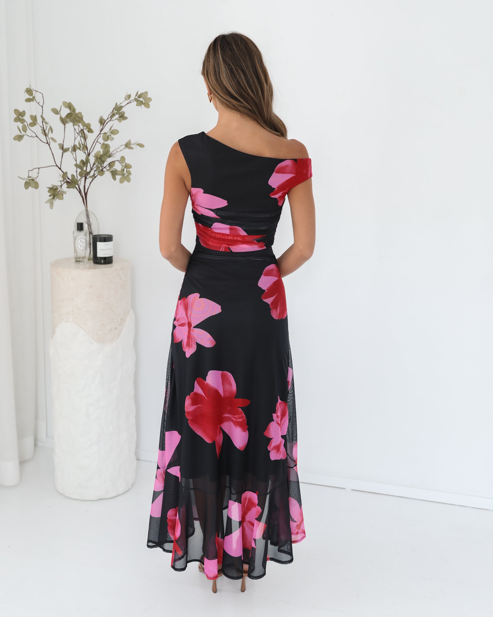 A woman standing in a white room wearing a black floral maxi dress with asymmetric neckline and ruffled skirt.