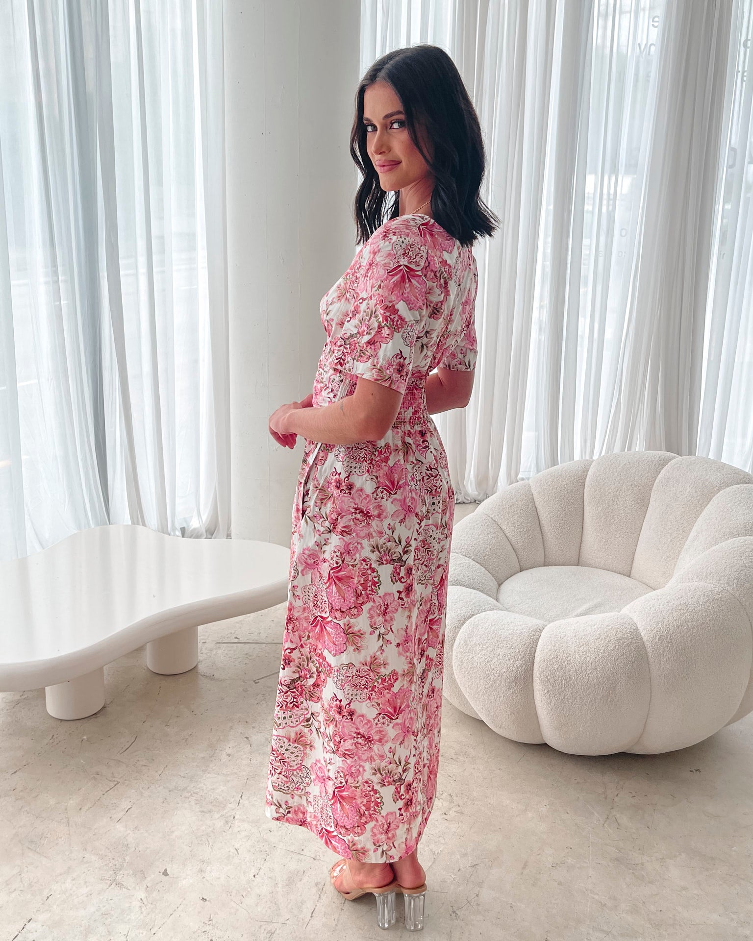 A woman standing in a room with a white chair, wearing a midi dress with a wide v-neckline, short puff sleeves, and an ornamental rose print.