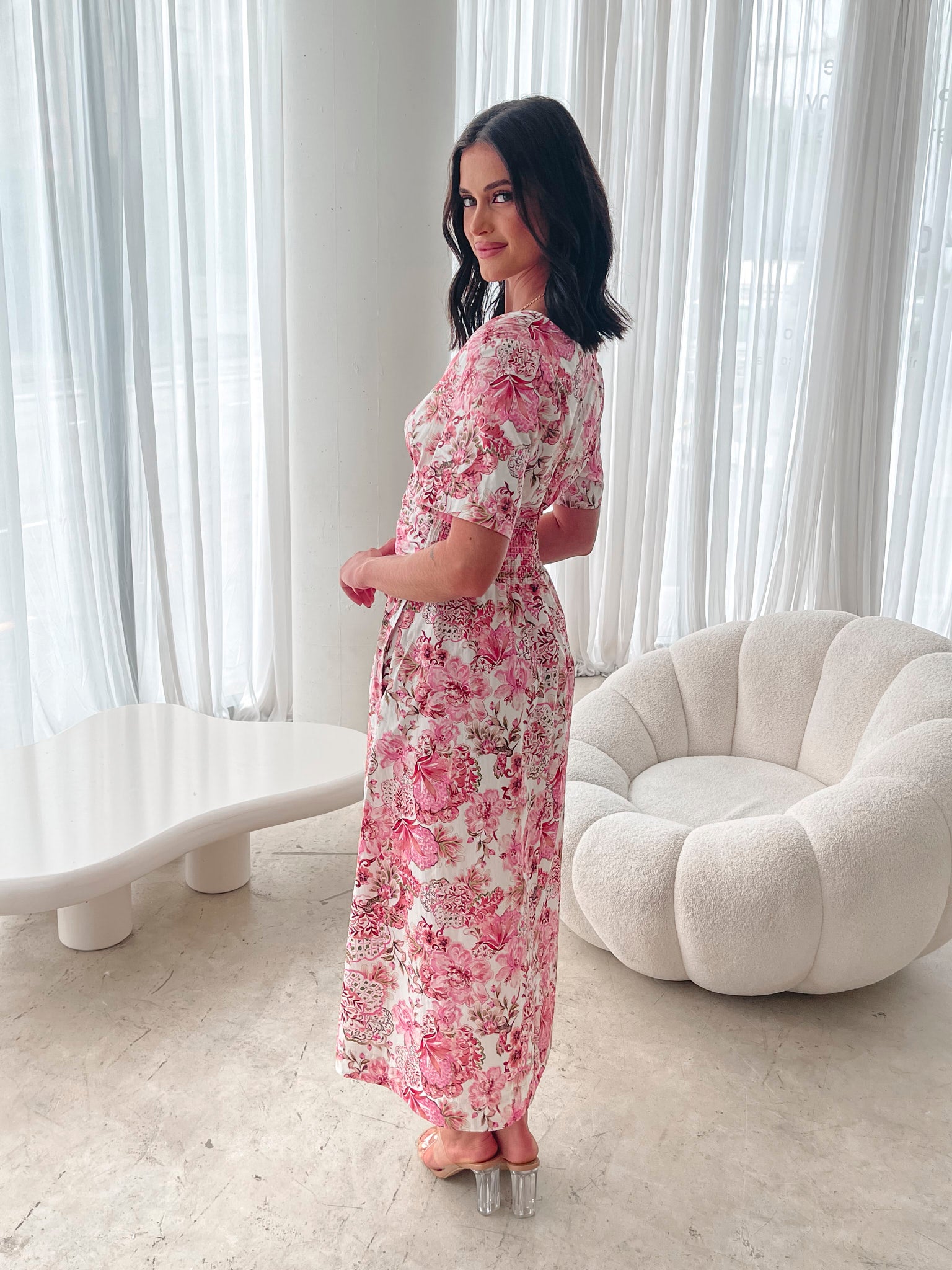 A woman standing in a room with a white chair, wearing a midi dress with a wide v-neckline, short puff sleeves, and an ornamental rose print.