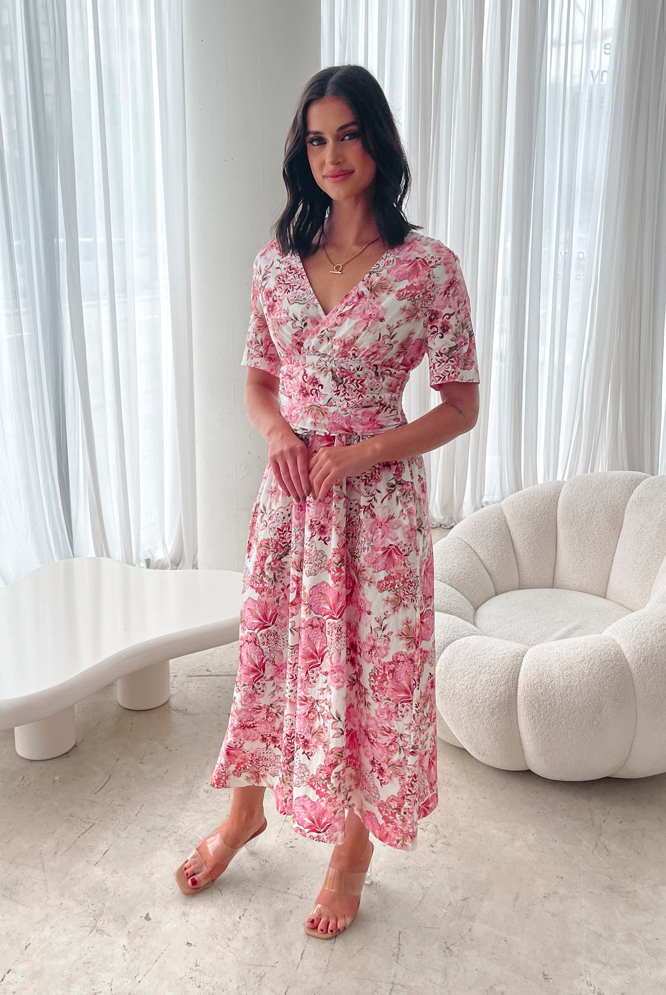 A woman standing in a room with a white chair, wearing a midi dress with a wide v-neckline, short puff sleeves, and an ornamental rose print.