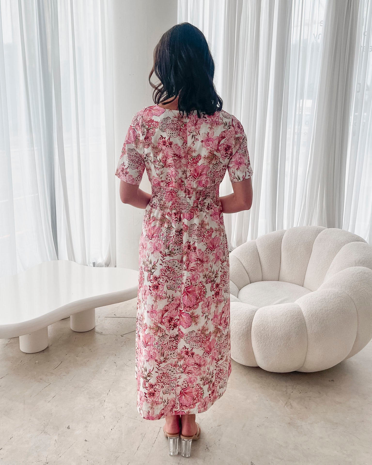 A woman standing in a room with a white chair, wearing a midi dress with a wide v-neckline, short puff sleeves, and an ornamental rose print.