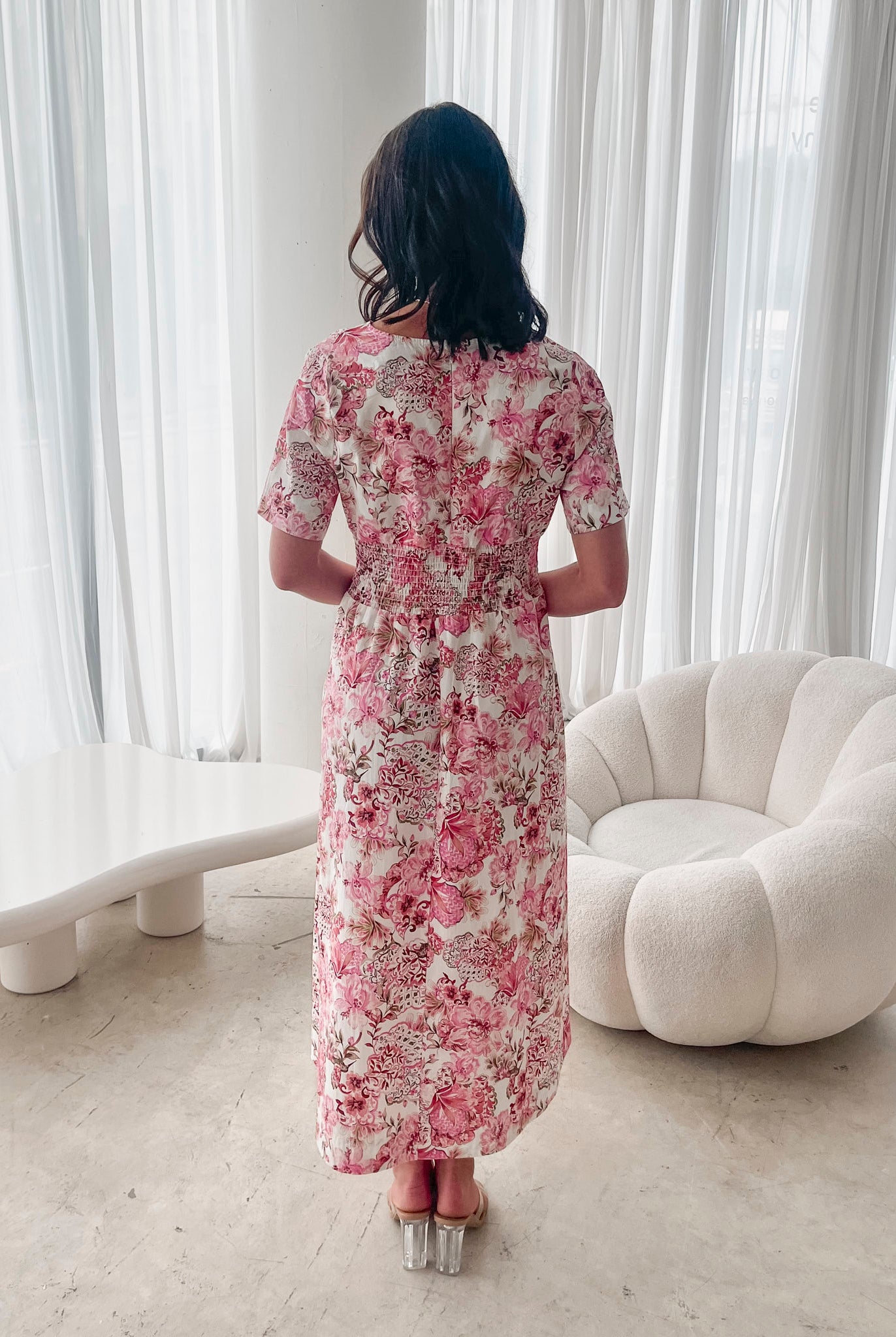 A woman standing in a room with a white chair, wearing a midi dress with a wide v-neckline, short puff sleeves, and an ornamental rose print.