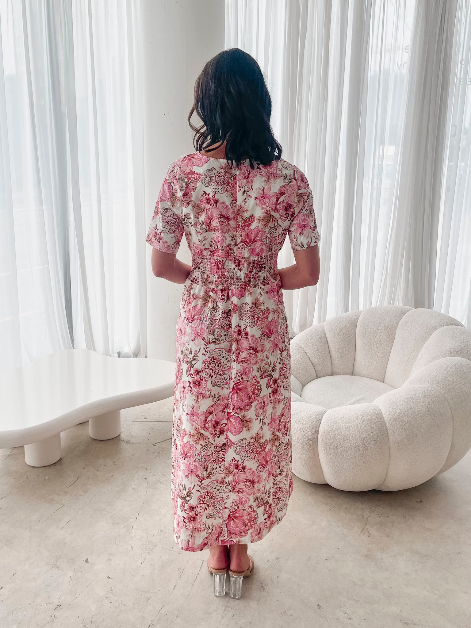 A woman standing in a room with a white chair, wearing a midi dress with a wide v-neckline, short puff sleeves, and an ornamental rose print.