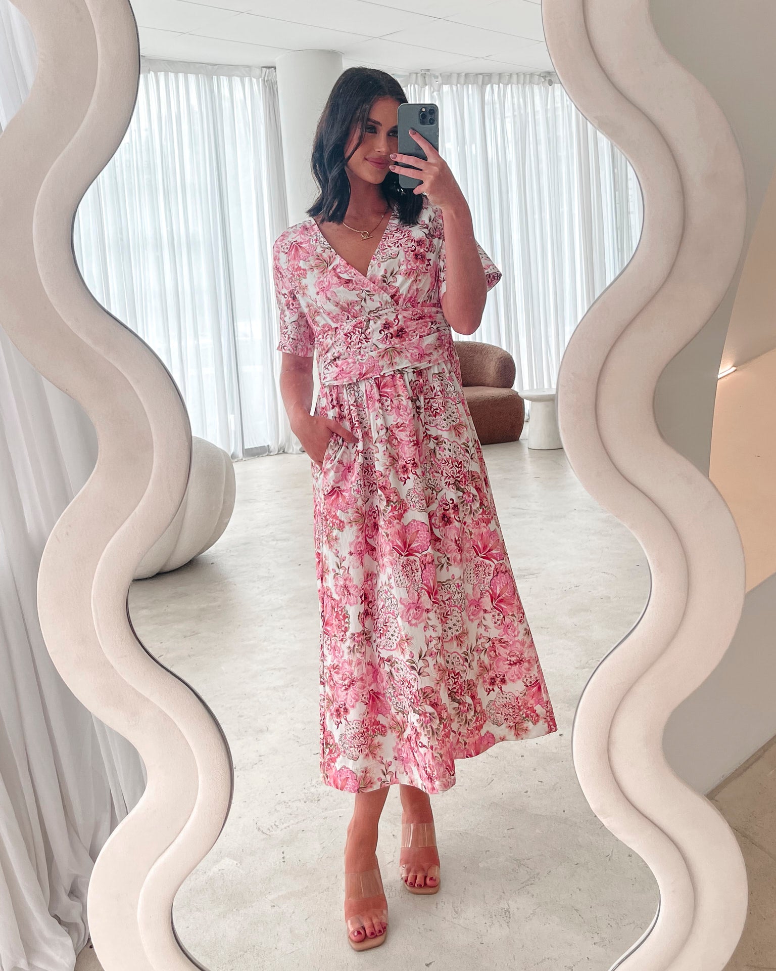 A woman standing in a room with a white chair, wearing a midi dress with a wide v-neckline, short puff sleeves, and an ornamental rose print.