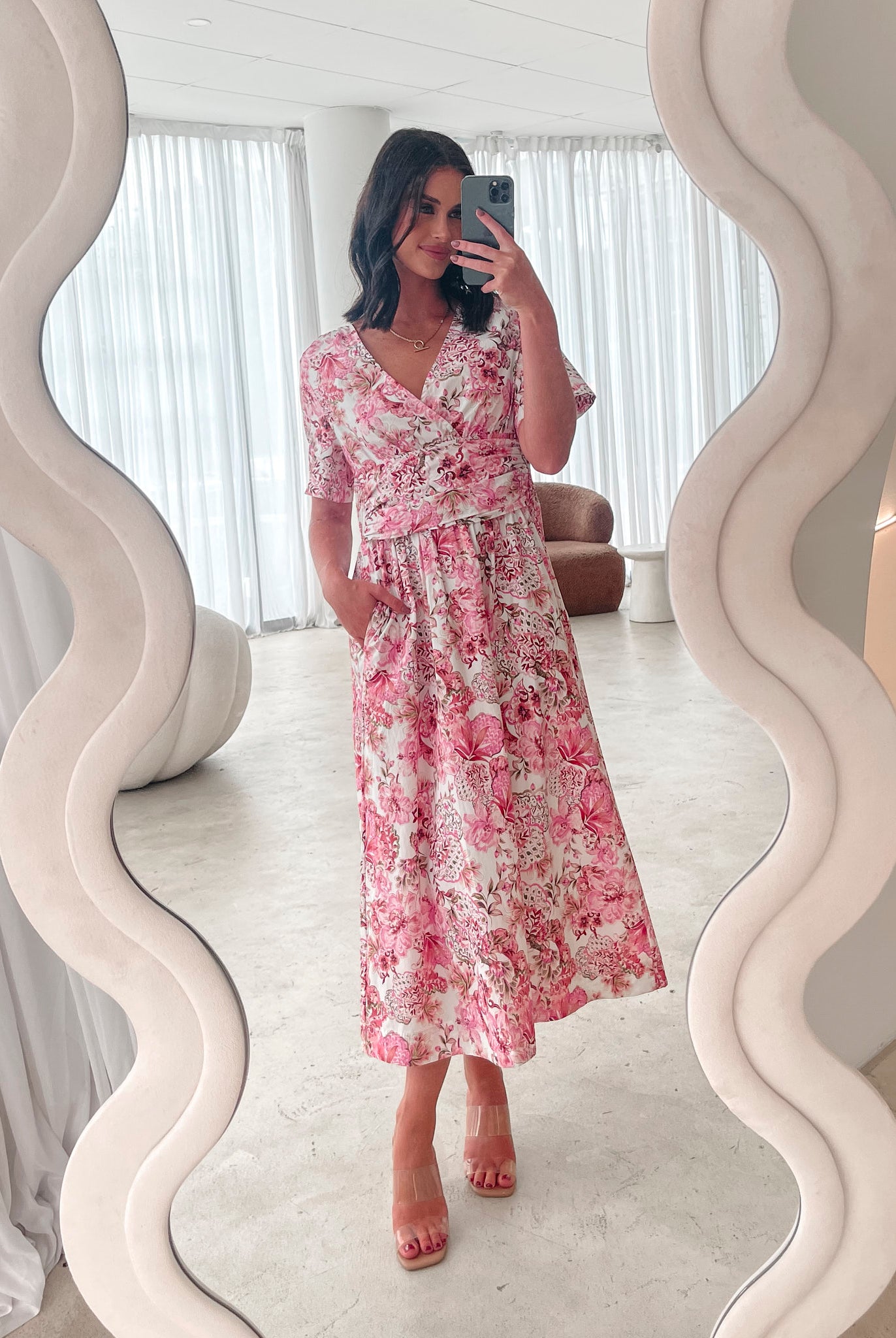 A woman standing in a room with a white chair, wearing a midi dress with a wide v-neckline, short puff sleeves, and an ornamental rose print.