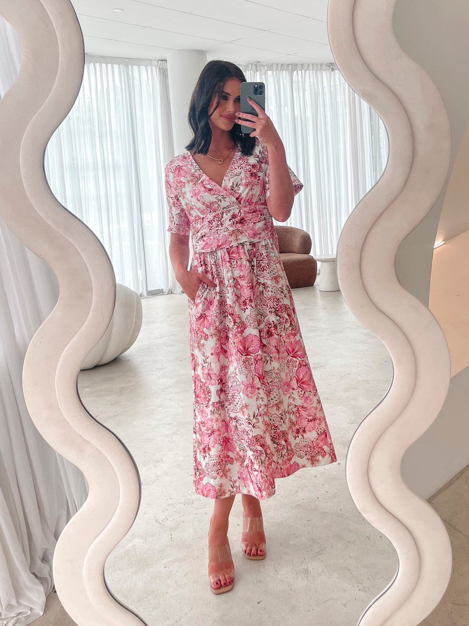 A woman standing in a room with a white chair, wearing a midi dress with a wide v-neckline, short puff sleeves, and an ornamental rose print.