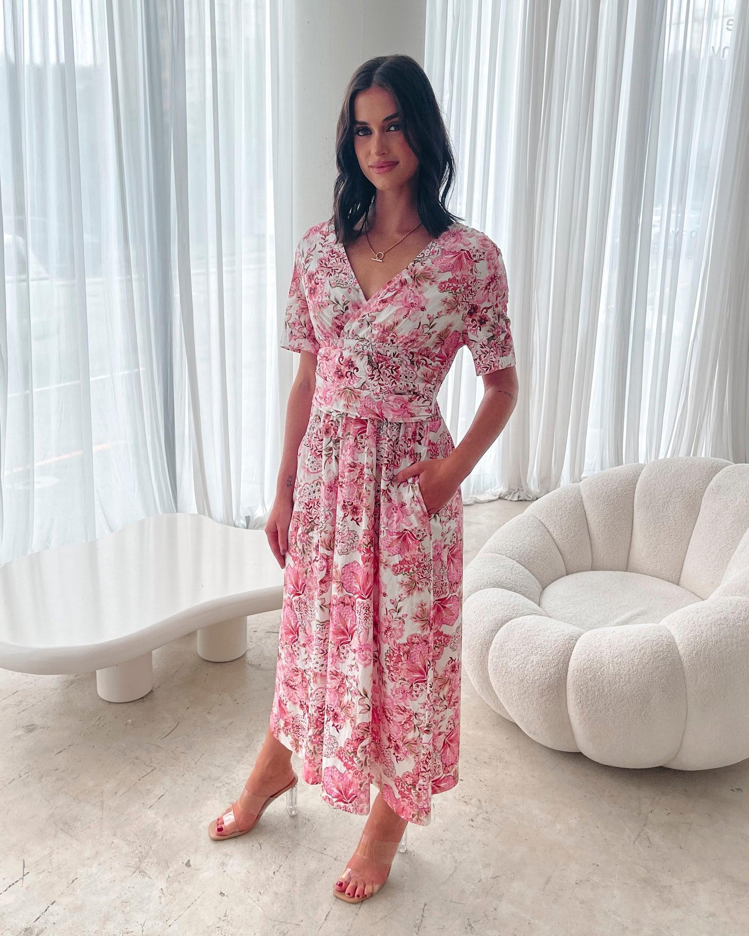 A woman standing in a room with a white chair, wearing a midi dress with a wide v-neckline, short puff sleeves, and an ornamental rose print.