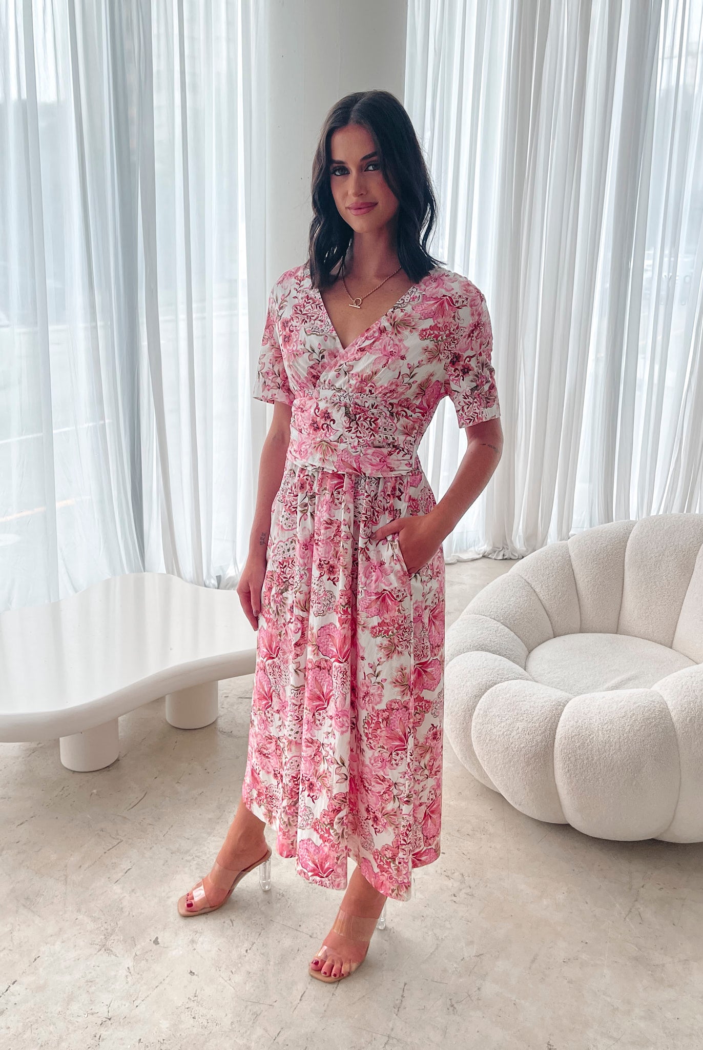 A woman standing in a room with a white chair, wearing a midi dress with a wide v-neckline, short puff sleeves, and an ornamental rose print.