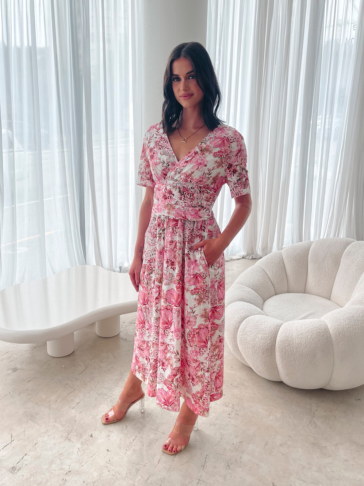 A woman standing in a room with a white chair, wearing a midi dress with a wide v-neckline, short puff sleeves, and an ornamental rose print.