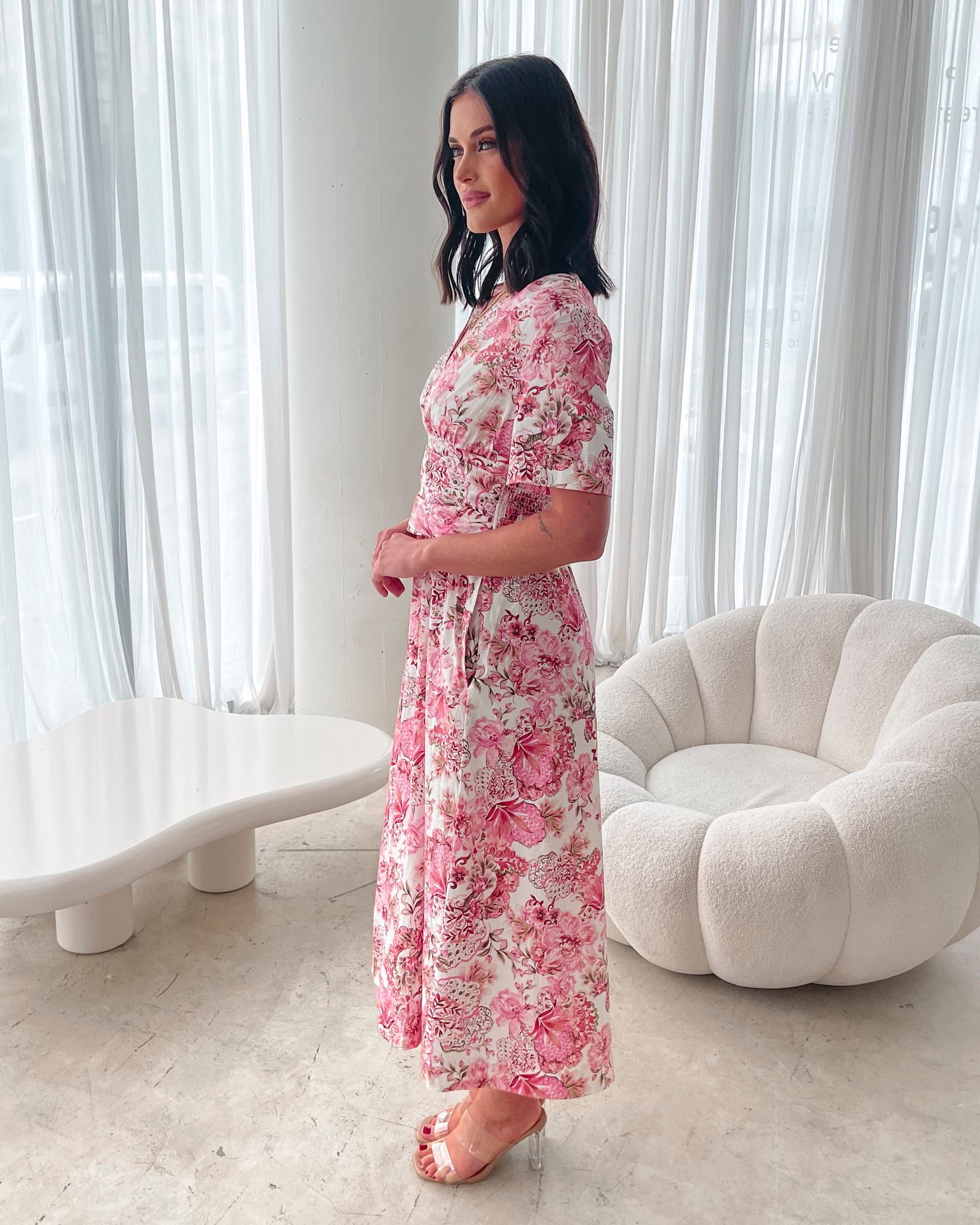 A woman standing in a room with a white chair, wearing a midi dress with a wide v-neckline, short puff sleeves, and an ornamental rose print.