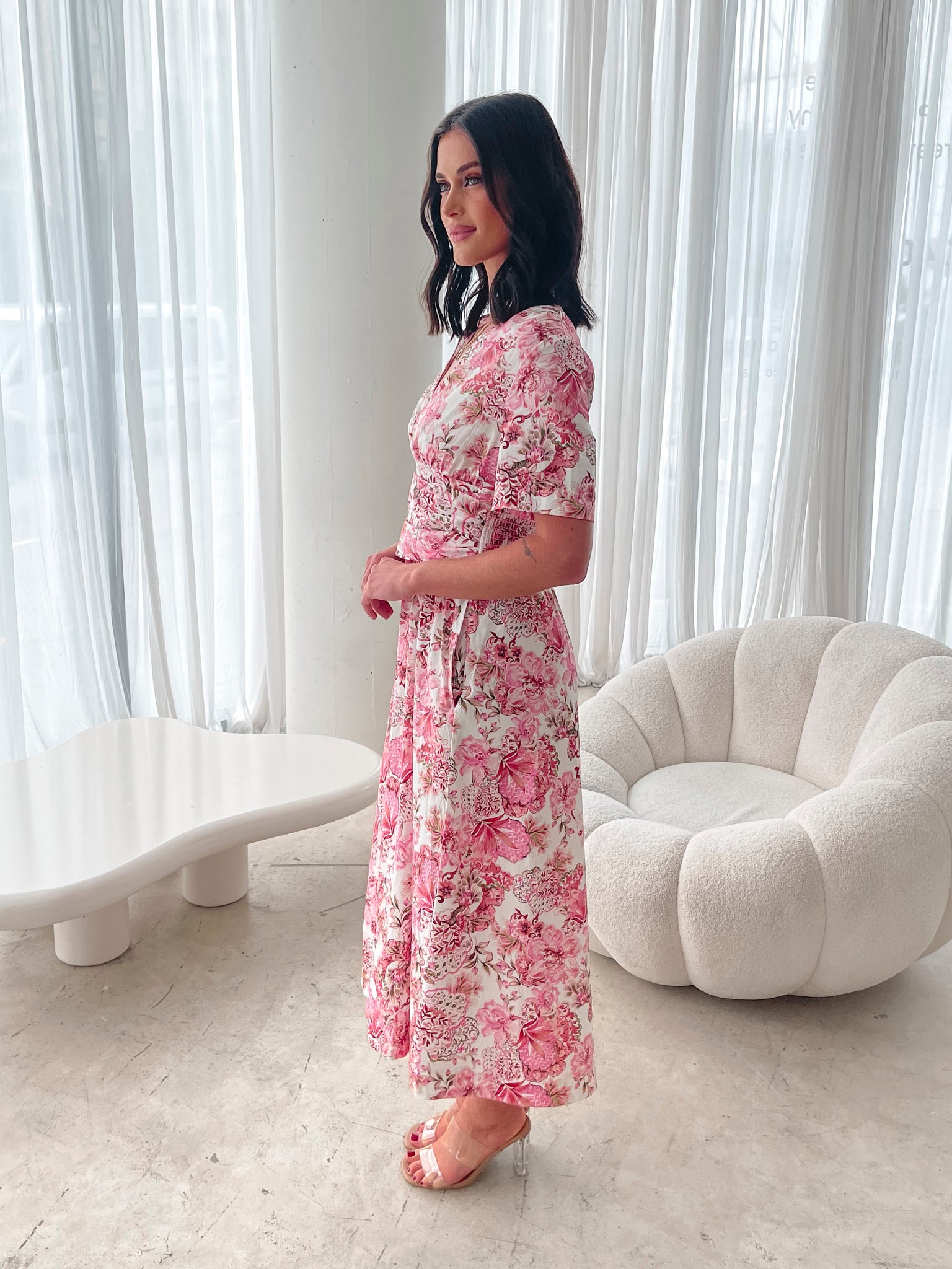 A woman standing in a room with a white chair, wearing a midi dress with a wide v-neckline, short puff sleeves, and an ornamental rose print.