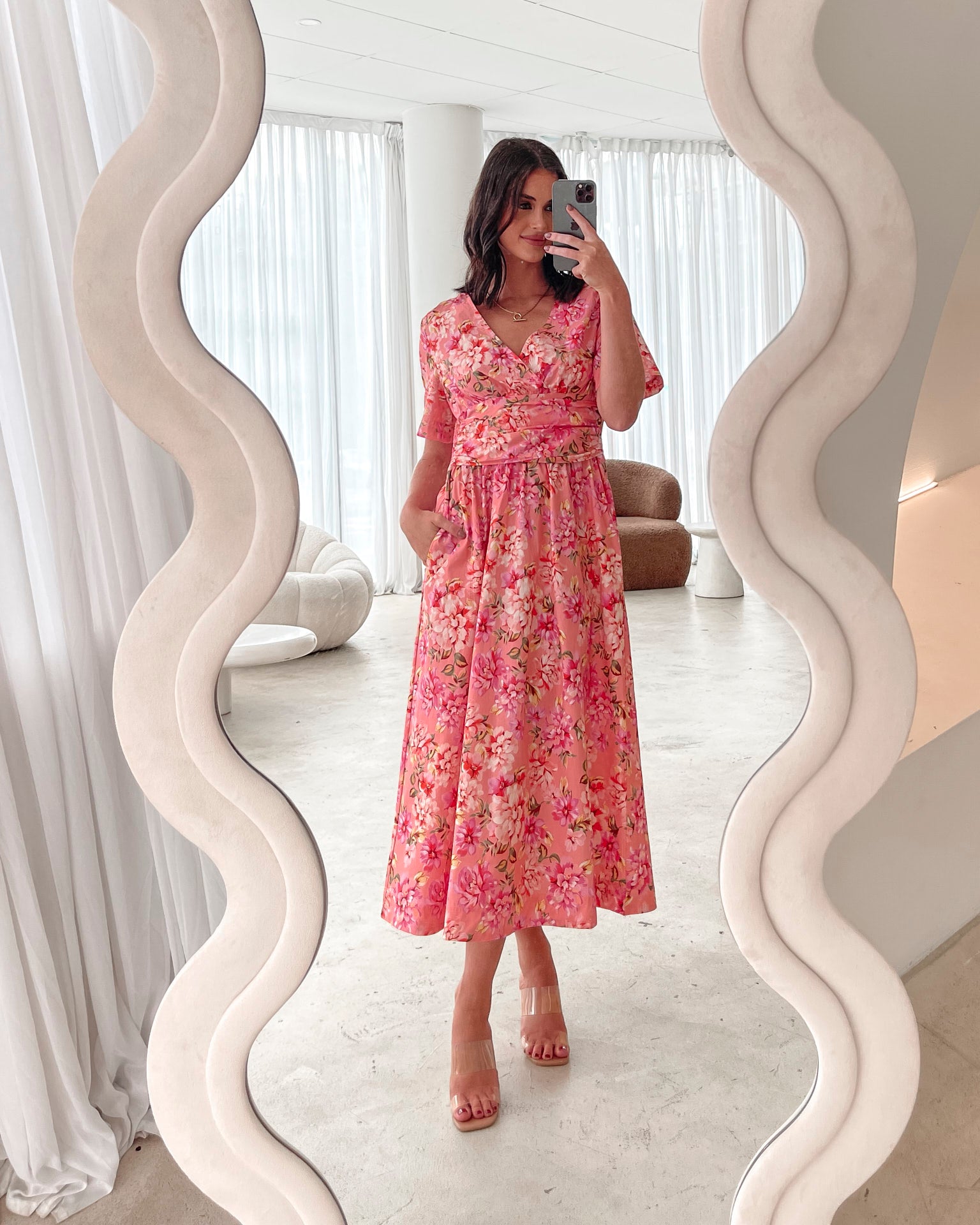 A person sitting on a white sofa wearing a peach blossom midi dress with a wide v-neckline, short puff sleeves, and a flowy a-line skirt.