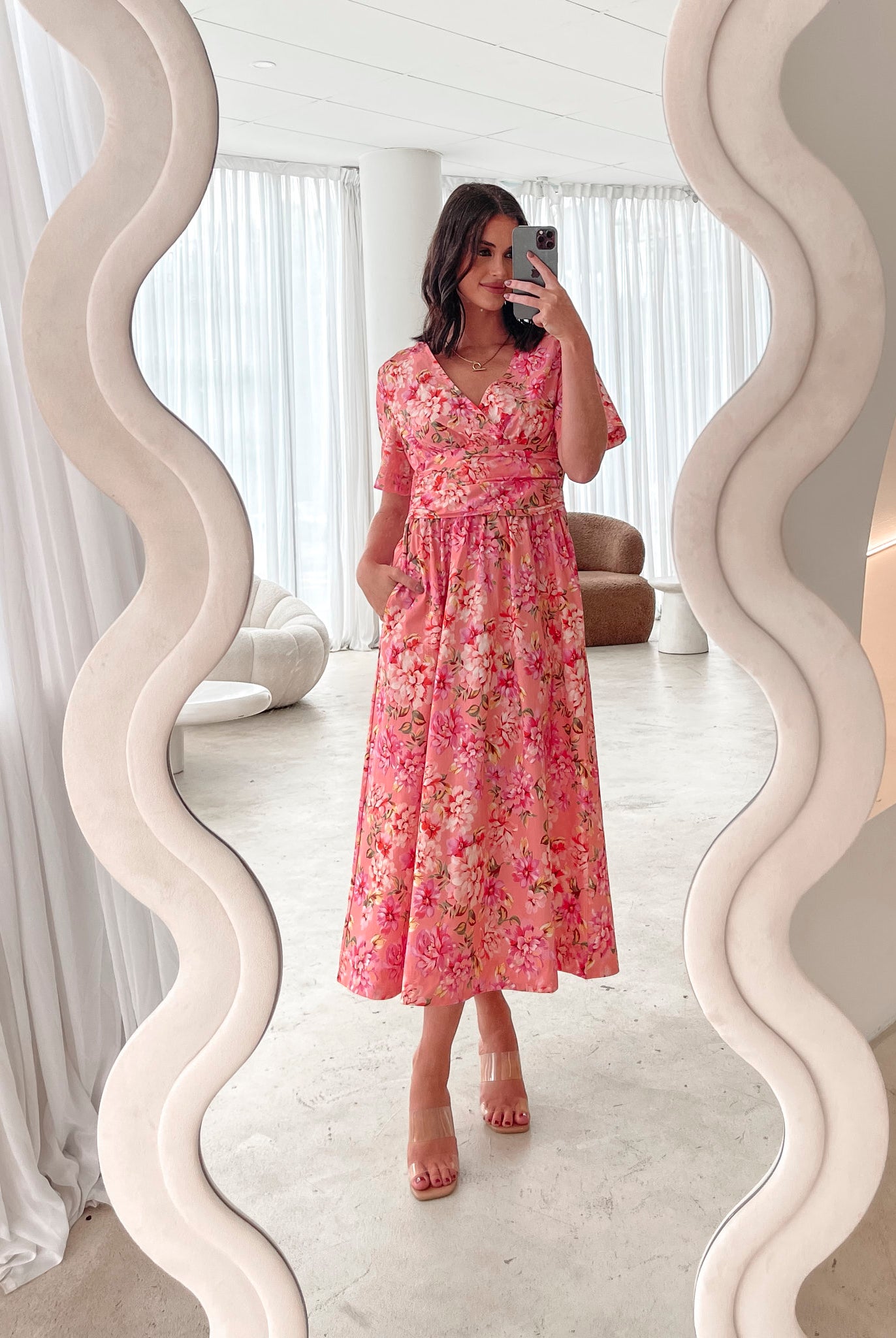 A person sitting on a white sofa wearing a peach blossom midi dress with a wide v-neckline, short puff sleeves, and a flowy a-line skirt.