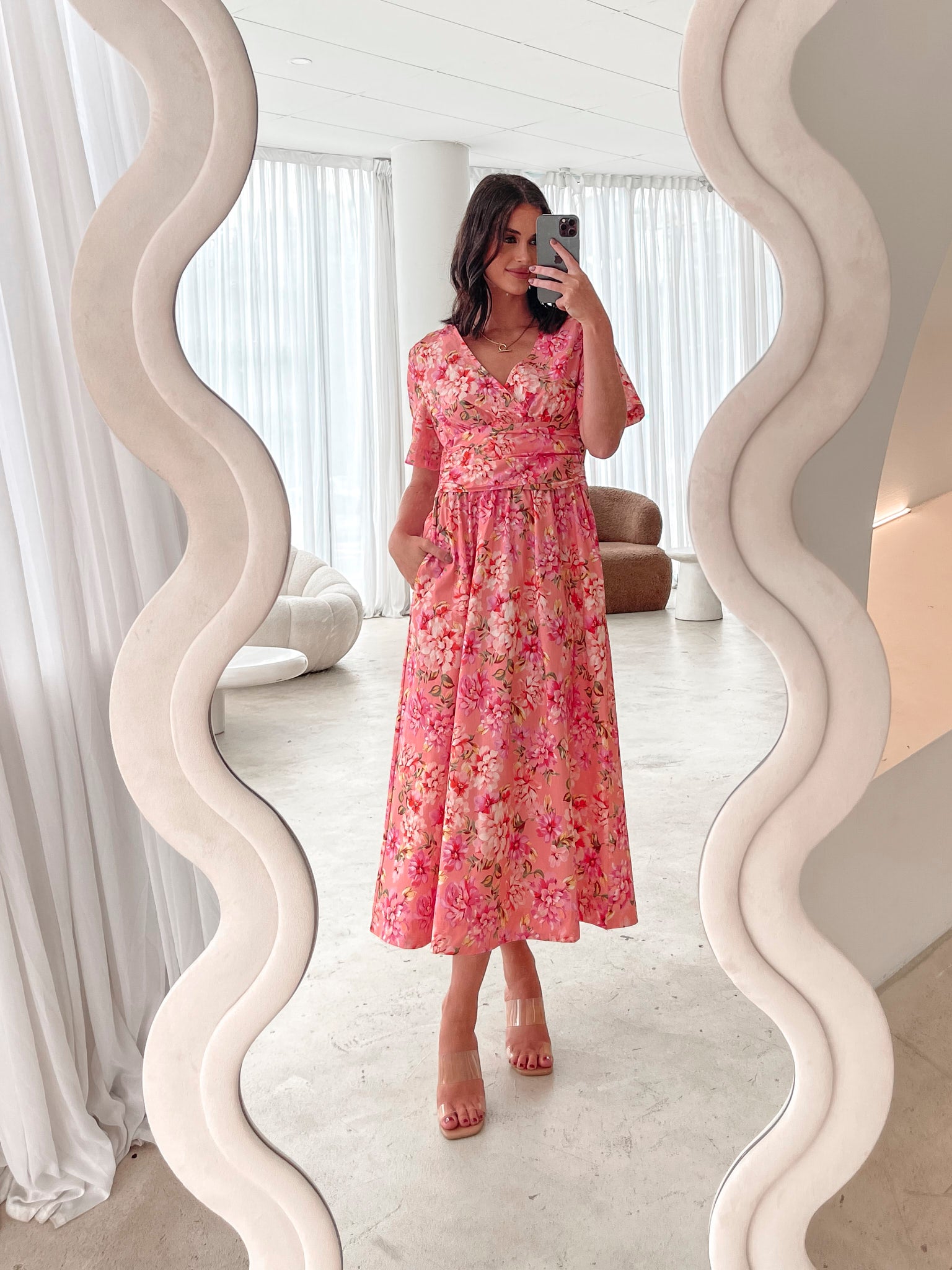 A person sitting on a white sofa wearing a peach blossom midi dress with a wide v-neckline, short puff sleeves, and a flowy a-line skirt.