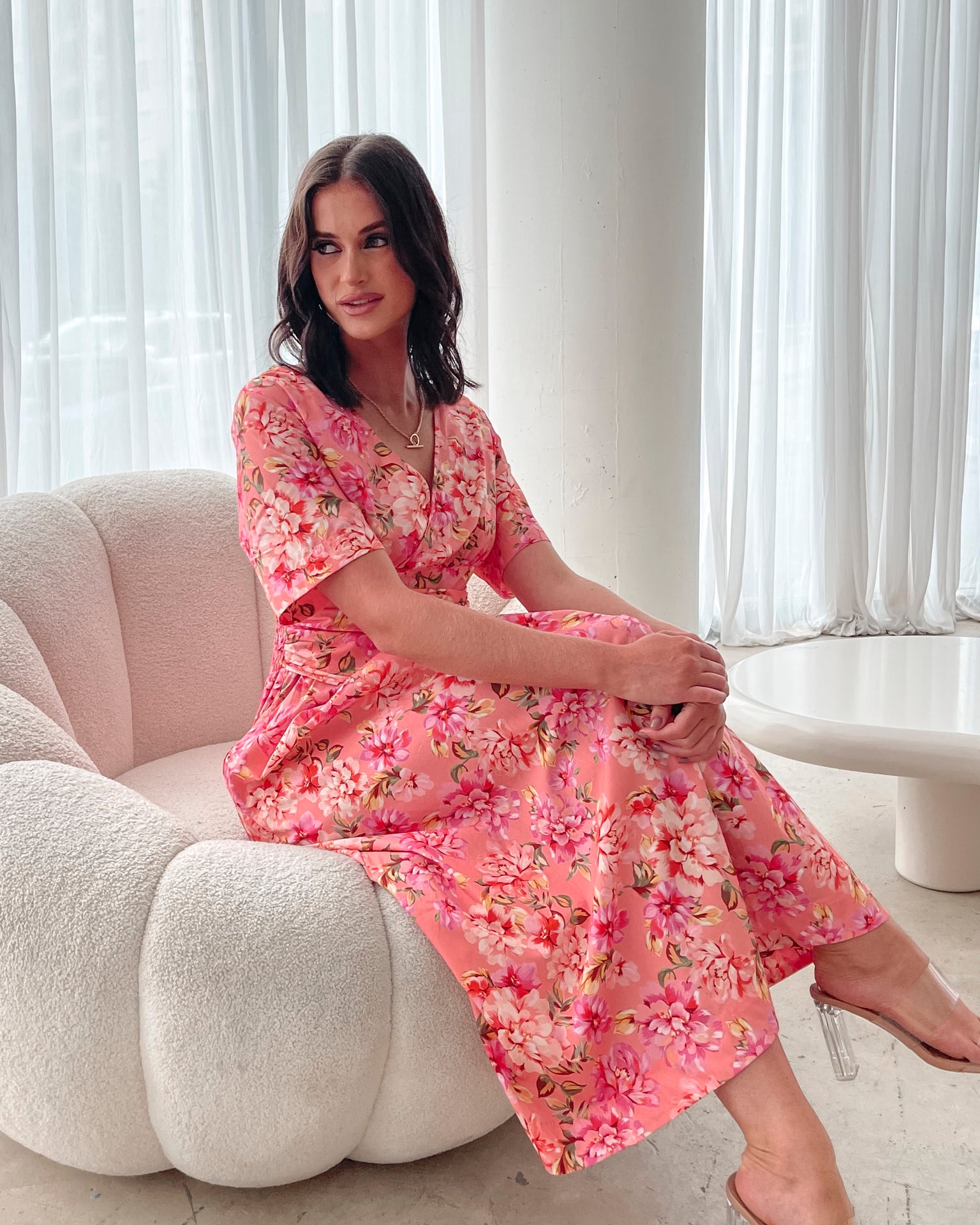 A person sitting on a white sofa wearing a peach blossom midi dress with a wide v-neckline, short puff sleeves, and a flowy a-line skirt.
