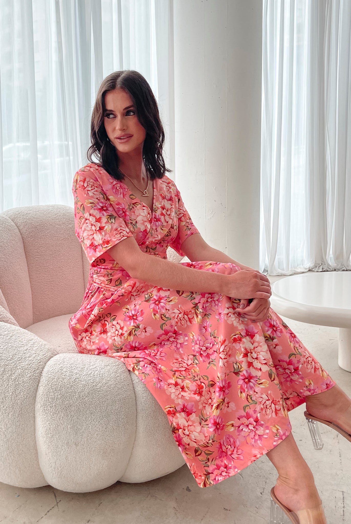 A person sitting on a white sofa wearing a peach blossom midi dress with a wide v-neckline, short puff sleeves, and a flowy a-line skirt.
