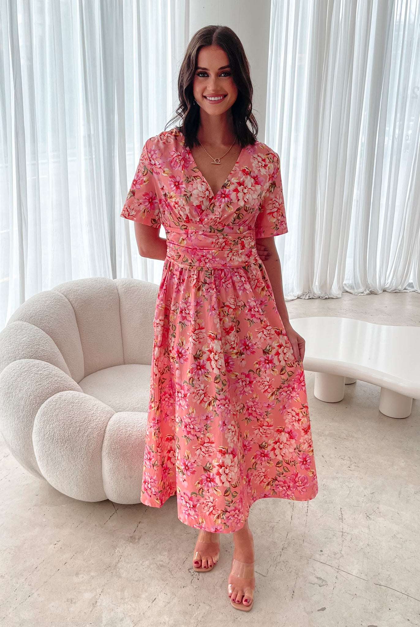 A person sitting on a white sofa wearing a peach blossom midi dress with a wide v-neckline, short puff sleeves, and a flowy a-line skirt.