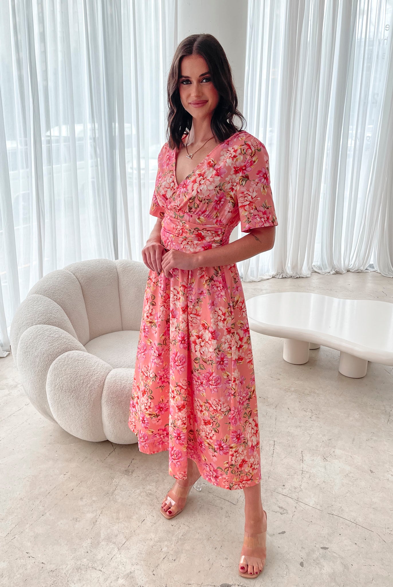A person sitting on a white sofa wearing a peach blossom midi dress with a wide v-neckline, short puff sleeves, and a flowy a-line skirt.