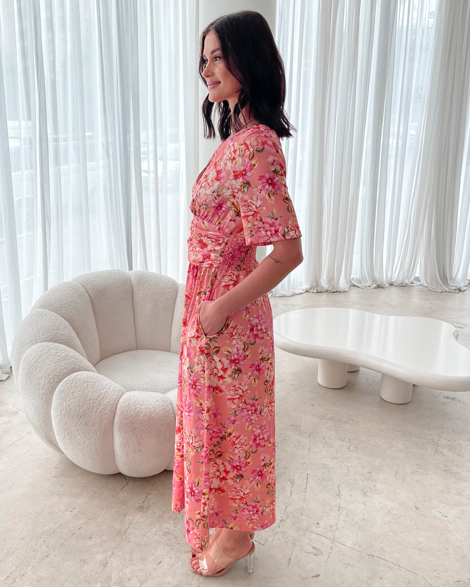 A person sitting on a white sofa wearing a peach blossom midi dress with a wide v-neckline, short puff sleeves, and a flowy a-line skirt.