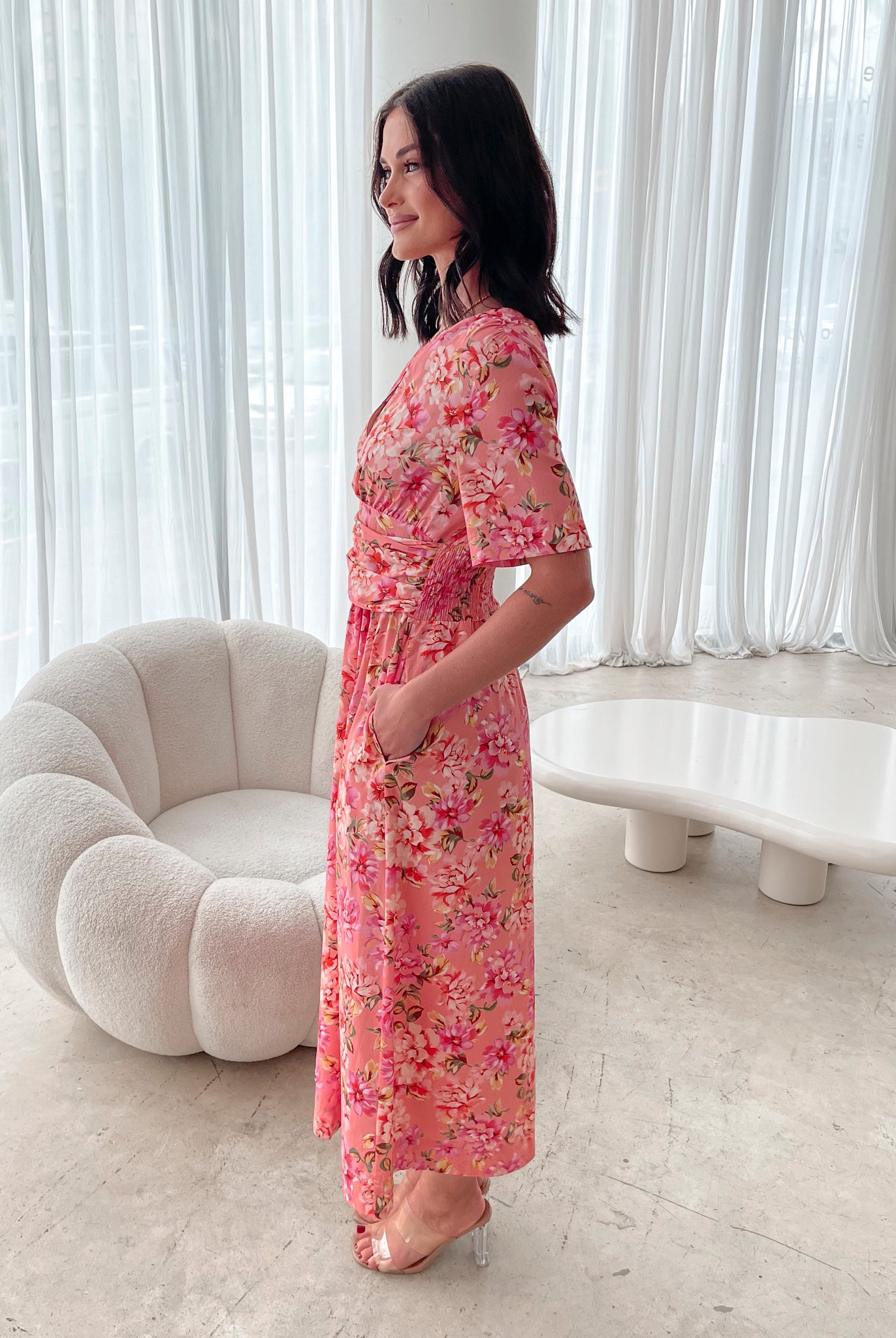 A person sitting on a white sofa wearing a peach blossom midi dress with a wide v-neckline, short puff sleeves, and a flowy a-line skirt.