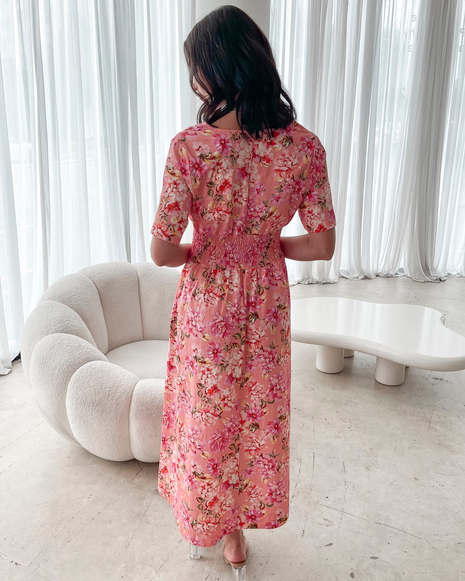 A person sitting on a white sofa wearing a peach blossom midi dress with a wide v-neckline, short puff sleeves, and a flowy a-line skirt.