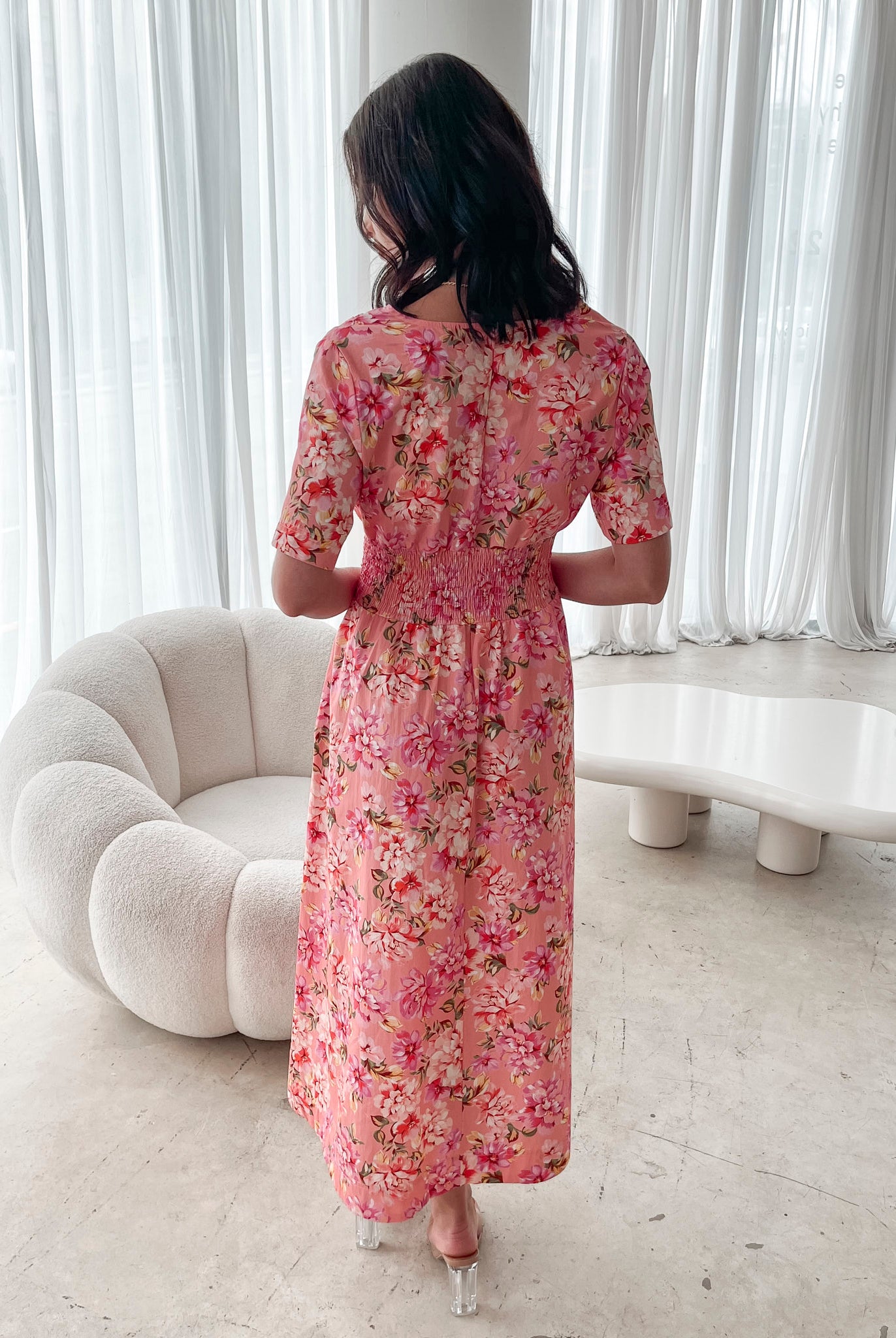 A person sitting on a white sofa wearing a peach blossom midi dress with a wide v-neckline, short puff sleeves, and a flowy a-line skirt.