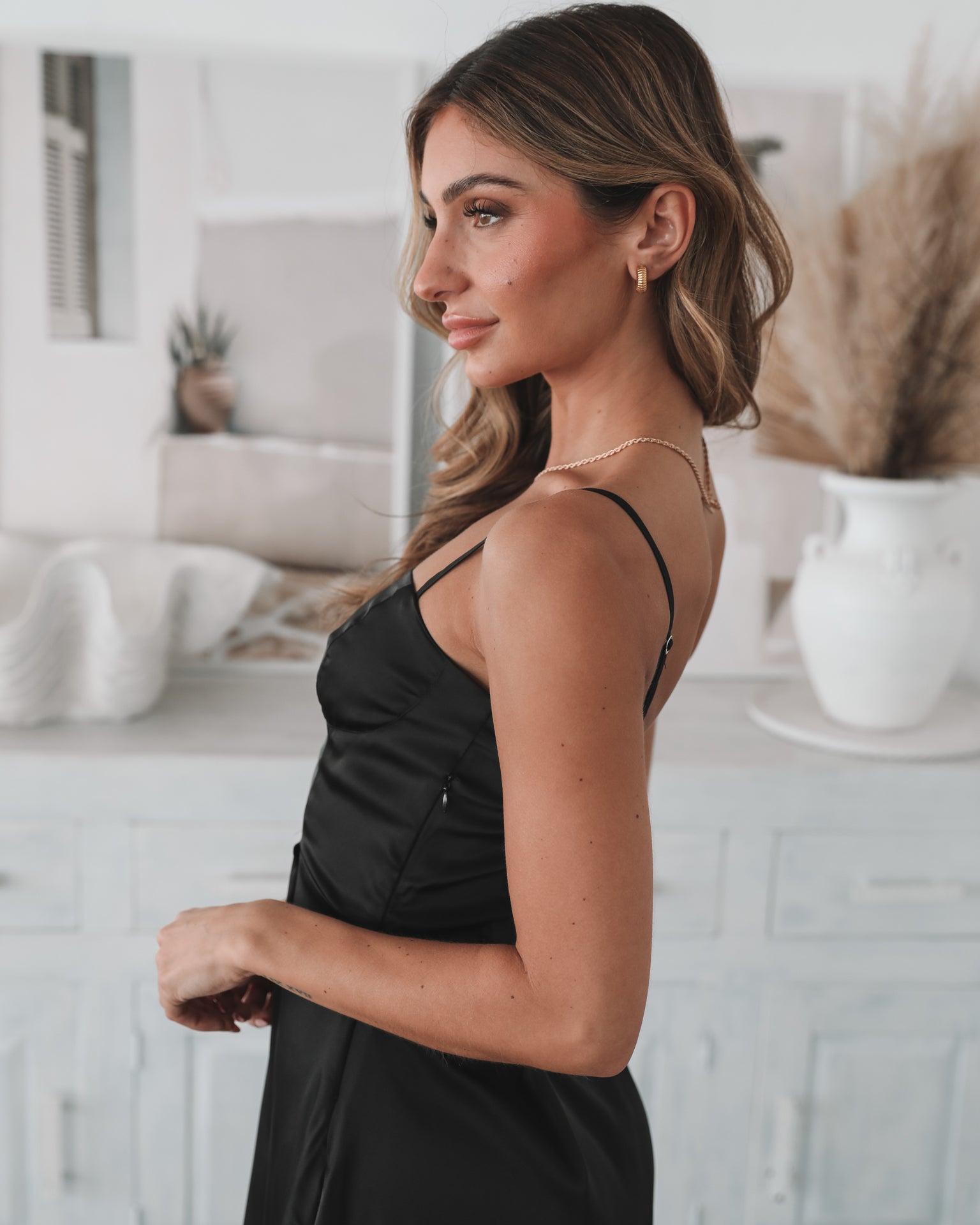 A woman standing in a room with light decor, wearing a black mini dress with a sweetheart neckline and slim straps. She has accessorized with a flower-shaped charm bracelet and is wearing block heels.