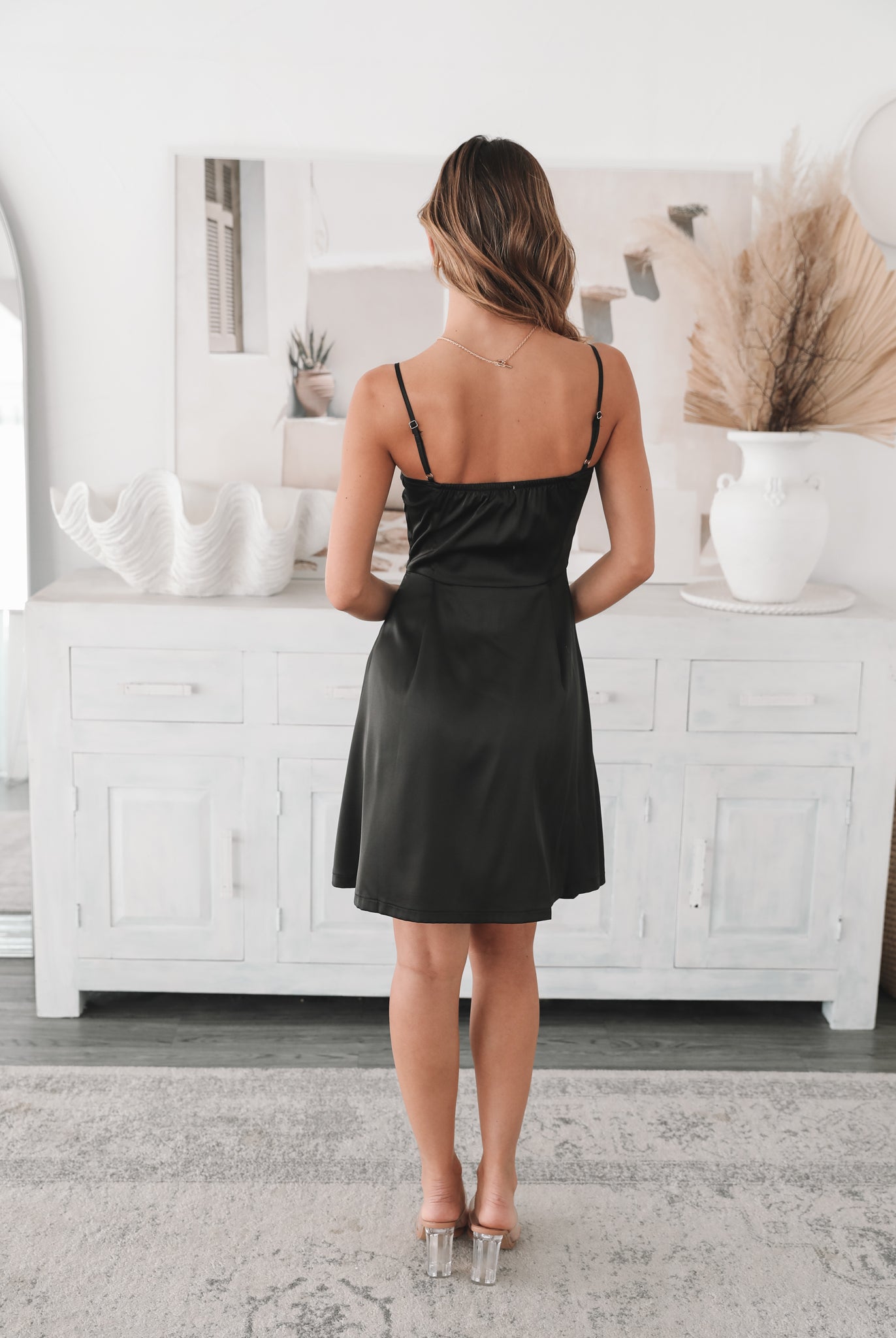 A woman standing in a room with light decor, wearing a black mini dress with a sweetheart neckline and slim straps. She has accessorized with a flower-shaped charm bracelet and is wearing block heels.