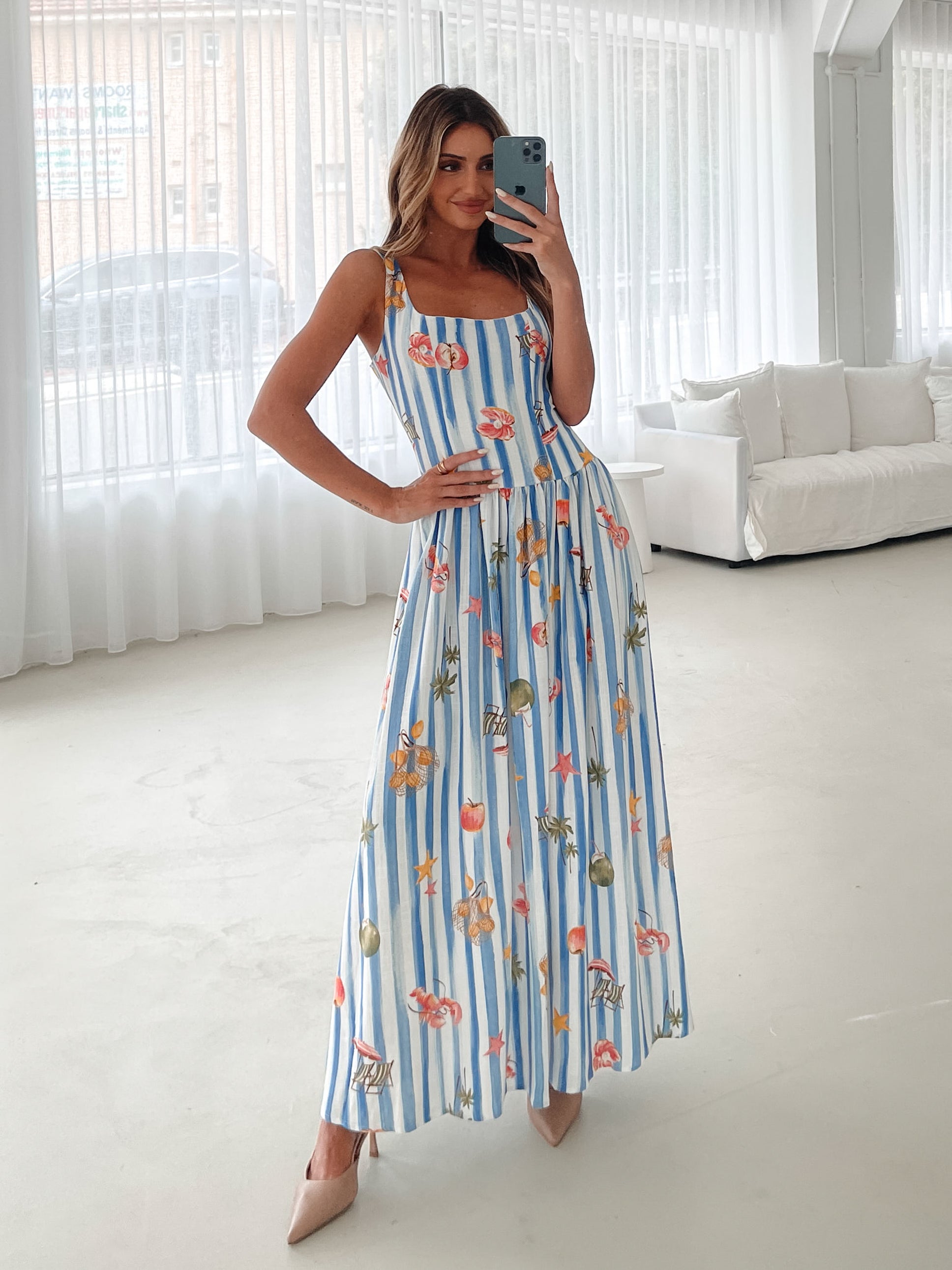 A model wearing a white and blue base multicolored print maxi dress with a square neckline and wide straps.